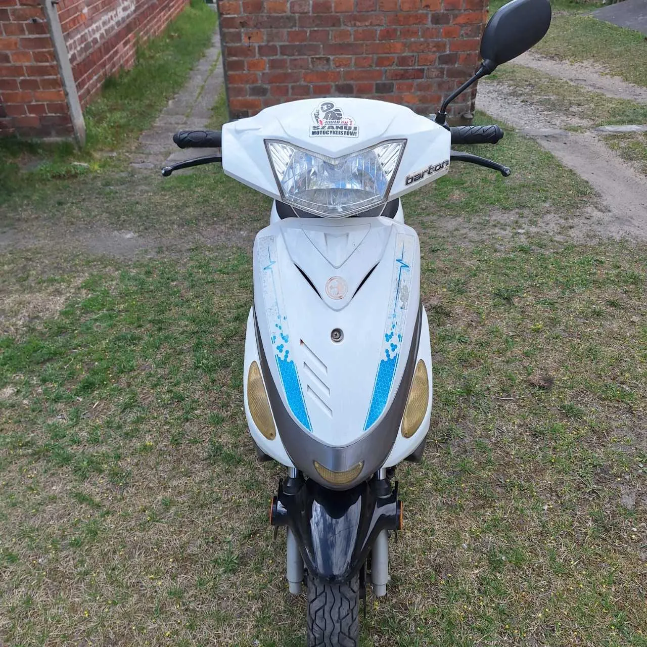 Biały skuter marki Barton z niebieskimi akcentami na przodzie, widok z przodu. Na osłonie przedniej naklejka z napisem "SZKOŁA MOTOCYKLISTÓW" oraz logo. Tło: ceglana ściana po lewej stronie, trawa z suchymi plamami i droga po prawej stronie. Skuter stoi na trawie.