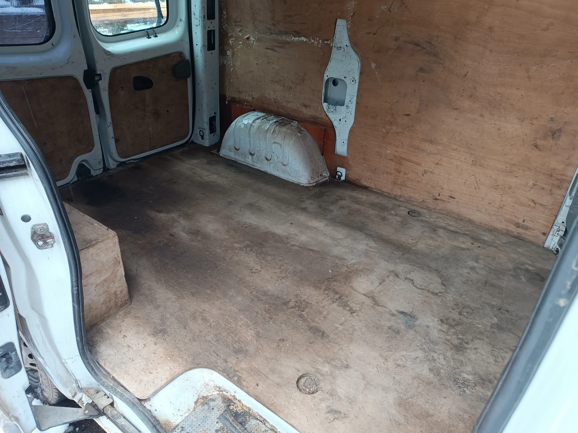 Wnętrze furgonetki z otwartymi tylnymi drzwiami. Podłoga jest brudna z widocznymi plamami i zadrapaniami. Po lewej stronie znajduje się drewniana skrzynia. Ściany wyłożone są płyta pilśniową, częściowo uszkodzoną. W prawej części widoczny jest metalowy element (prawdopodobnie osłona nadkola) oraz biały plastikowy uchwyt. Drzwi furgonetki mają okna i czarne uchwyty. Zewnętrzna część drzwi jest biała, z widocznymi śladami zużycia.