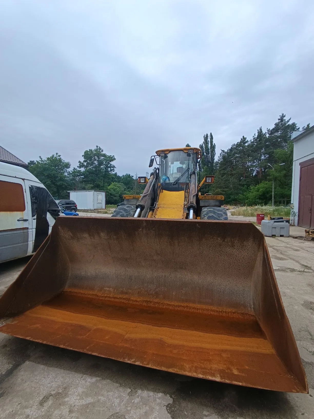 Zdjęcie przedstawia żółty ładowacz przedni z dużym, zardzewiałym metalowym łyżnikiem znajdującym się na pierwszym planie. Maszyna stoi na betonowym placu. W tle widoczny jest biały furgon po lewej stronie, mały biały budynek gospodarczy oraz pas zielonych drzew pod zachmurzonym niebem.
