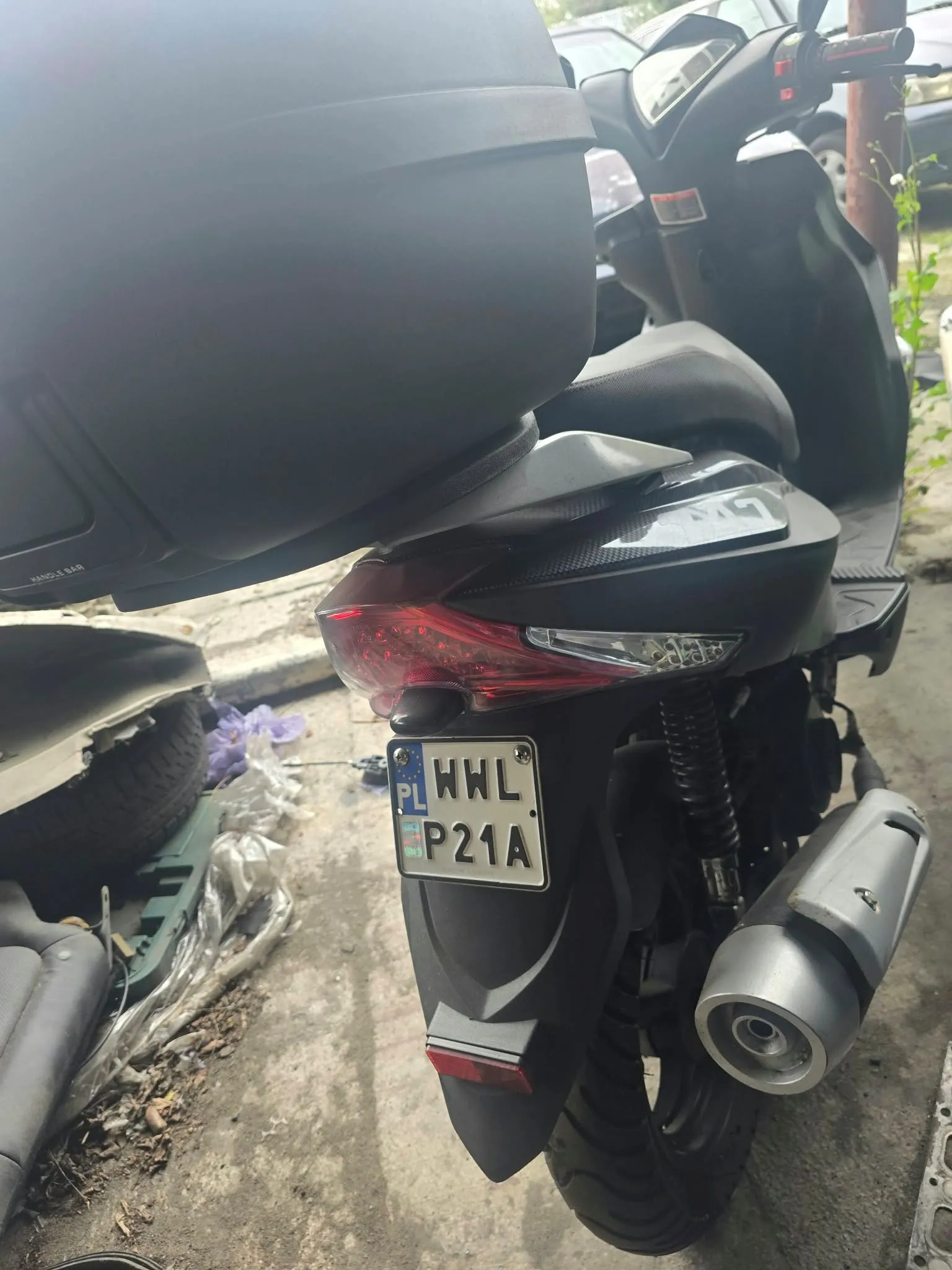 Zbliżenie na tył ciemnoszarego skutera z zamontowanym dużym, czarnym kufrem. Widoczna jest tylna lampa oraz polska tablica rejestracyjna o numerze WHL P21A. Po prawej stronie znajduje się srebrny tłumik wydechu. W tle po lewej widoczny jest fragment innego pojazdu i folia ochronna na ziemi.