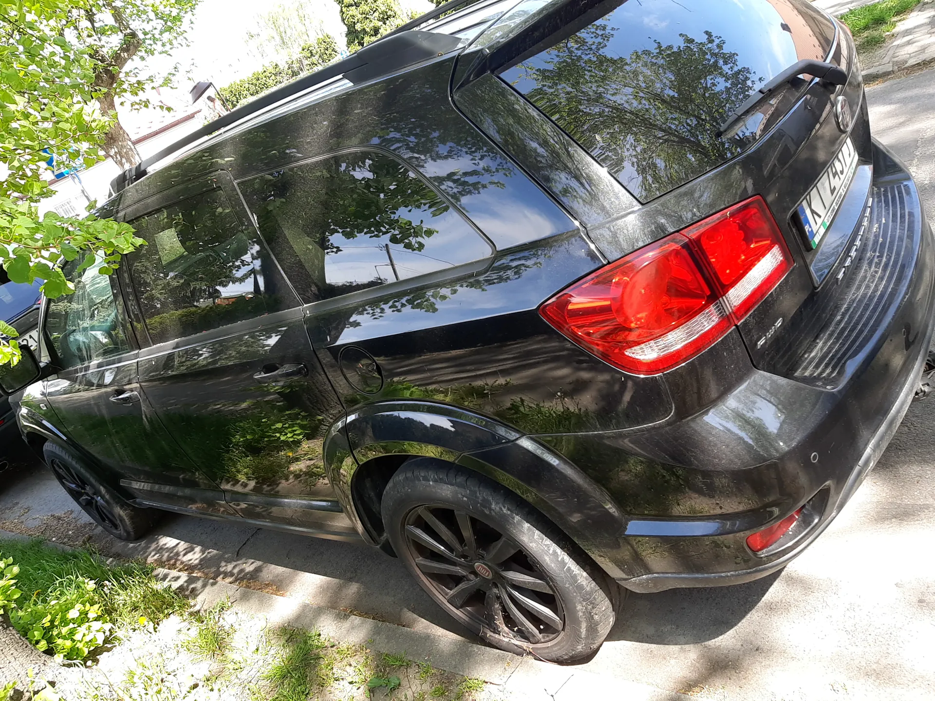 Czarny samochód typu SUV zaparkowany na poboczu. Widok z tyłu i boku. Widoczne czerwone światła tylne, tablica rejestracyjna KT 2491J, czarne felgi. W tle drzewa, zieleń i budynki. Słoneczna pogoda.