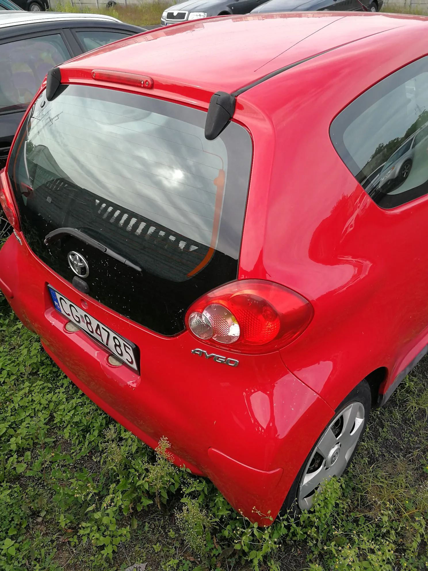 Tylna część czerwonego samochodu marki Chevrolet Aveo. Widoczna jest tylna szyba z wycieraczką, prawe tylne światło oraz tablica rejestracyjna o numerze CG 84785. Samochód stoi na trawiastym podłożu.
