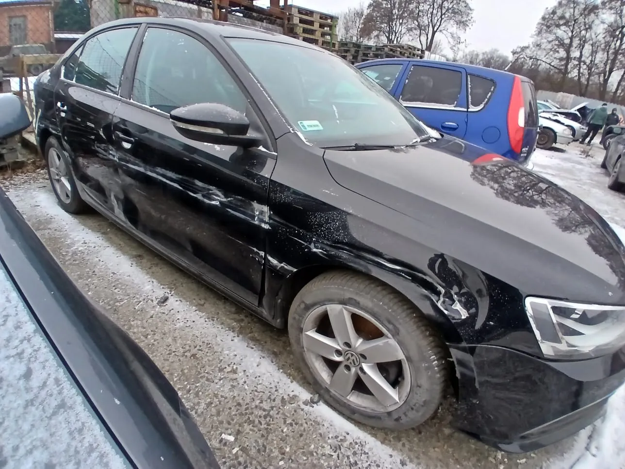 Czarny samochód osobowy marki Volkswagen z uszkodzeniami na przedniej prawej stronie, zaparkowany na nawierzchni z resztkami śniegu i żwirem. W tle niebieski samochód, inne pojazdy, budynki oraz drzewa bez liści. Na przedniej szybie widoczny mały zielony znaczek. Opony i felgi w stanie zużycia.