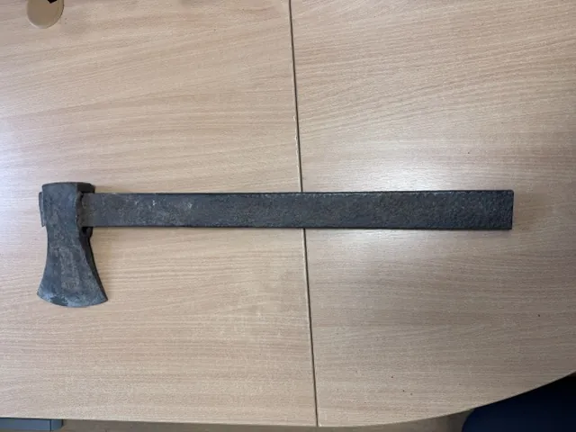 Stary, zardzewiały topór z metalową głową i długim, prostym metalowym trzonkiem, leżący poziomo na jasnobrązowym blacie stołu o strukturze drewna.