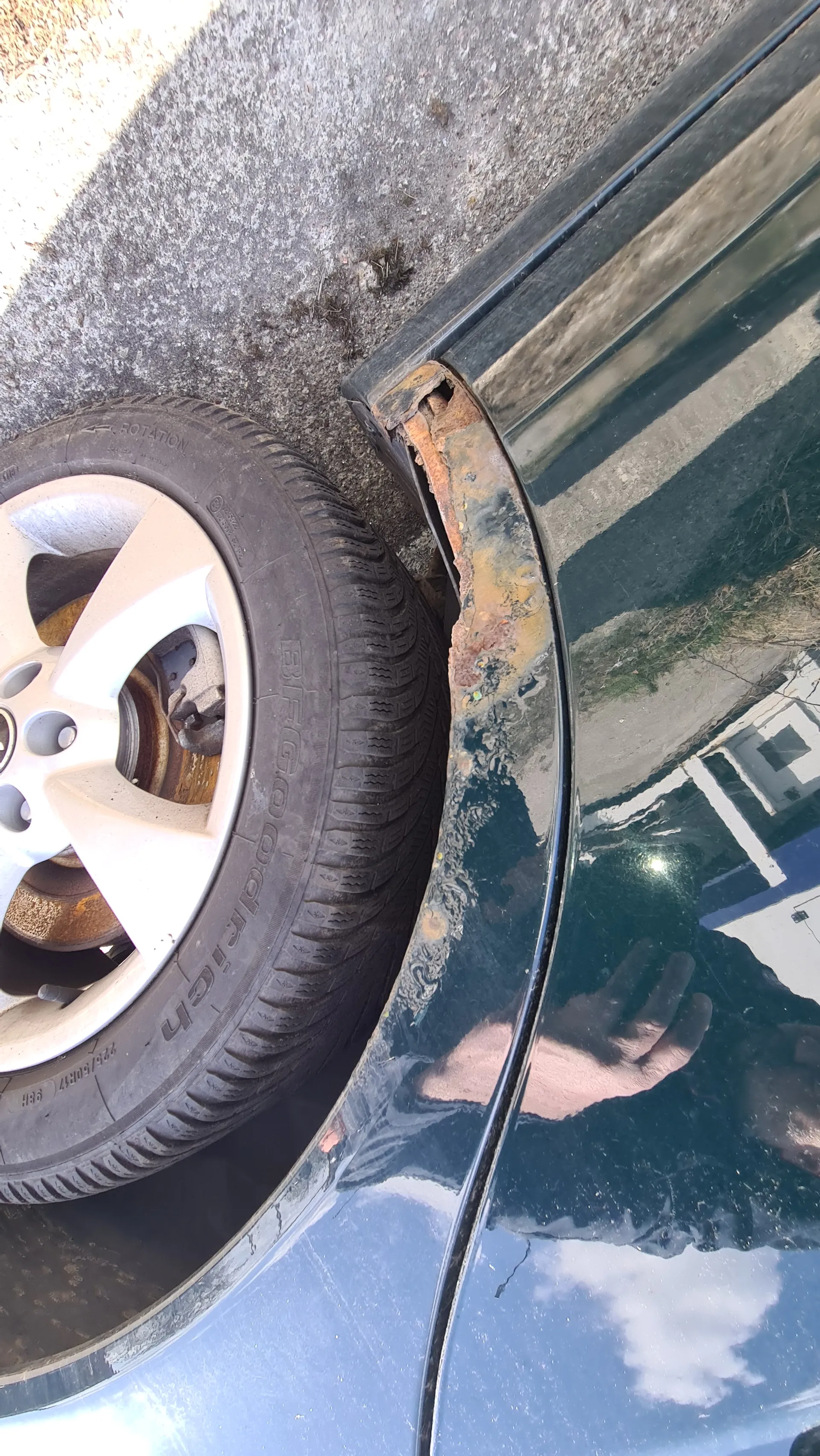 Zbliżenie na koło samochodu z oponą marki BFGoodrich. Widoczna rdza na nadkolu, srebrna felga i betonowa powierzchnia pod samochodem. W odbiciu na karoserii widać osobę trzymającą telefon, budynek z białymi i niebieskimi elementami oraz niebo z chmurami.