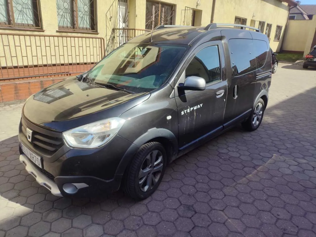 Ciemnoszary samochód dostawczy marki Dacia Stepway zaparkowany na brukowanej powierzchni. Na boku pojazdu widoczny napis "STEPWAY". Przedni zderzak z białym elementem, tablica rejestracyjna z częściowo widocznym numerem. W tle żółty budynek z oknami o metalowych kratkach i bramą. Podłoga wyłożona sześciokątnymi płytami brukowymi.