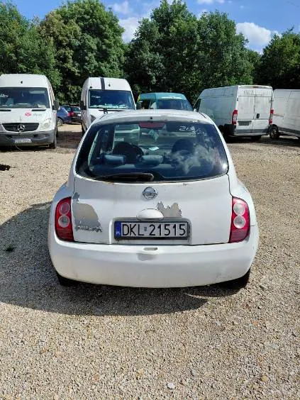 Widok z tyłu białego samochodu osobowego marki Nissan zaparkowanego na żwirowym placu. Samochód ma polską tablicę rejestracyjną o numerze DKL 21515. Na klapie bagażnika widoczne są uszkodzenia lakieru. W tle znajdują się inne białe samochody dostawcze oraz zielone drzewa pod niebieskim niebem.