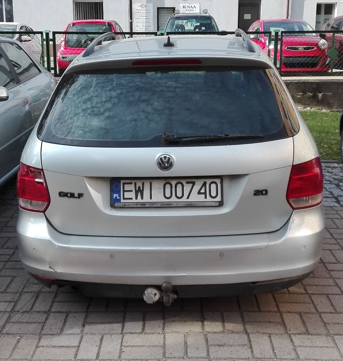 Srebrny samochód marki Volkswagen Golf w wersji kombi, zaparkowany tyłem do obiektywu. Na samochodzie widoczna jest polska tablica rejestracyjna o numerze EWI 00740 oraz napis GOLF po lewej stronie i 20 po prawej stronie klapy bagażnika. Pojazd posiada zamontowany hak holowniczy. W tle widoczne są inne zaparkowane samochody, w tym dwa czerwone, oraz zielony płot i fragmenty budynków.