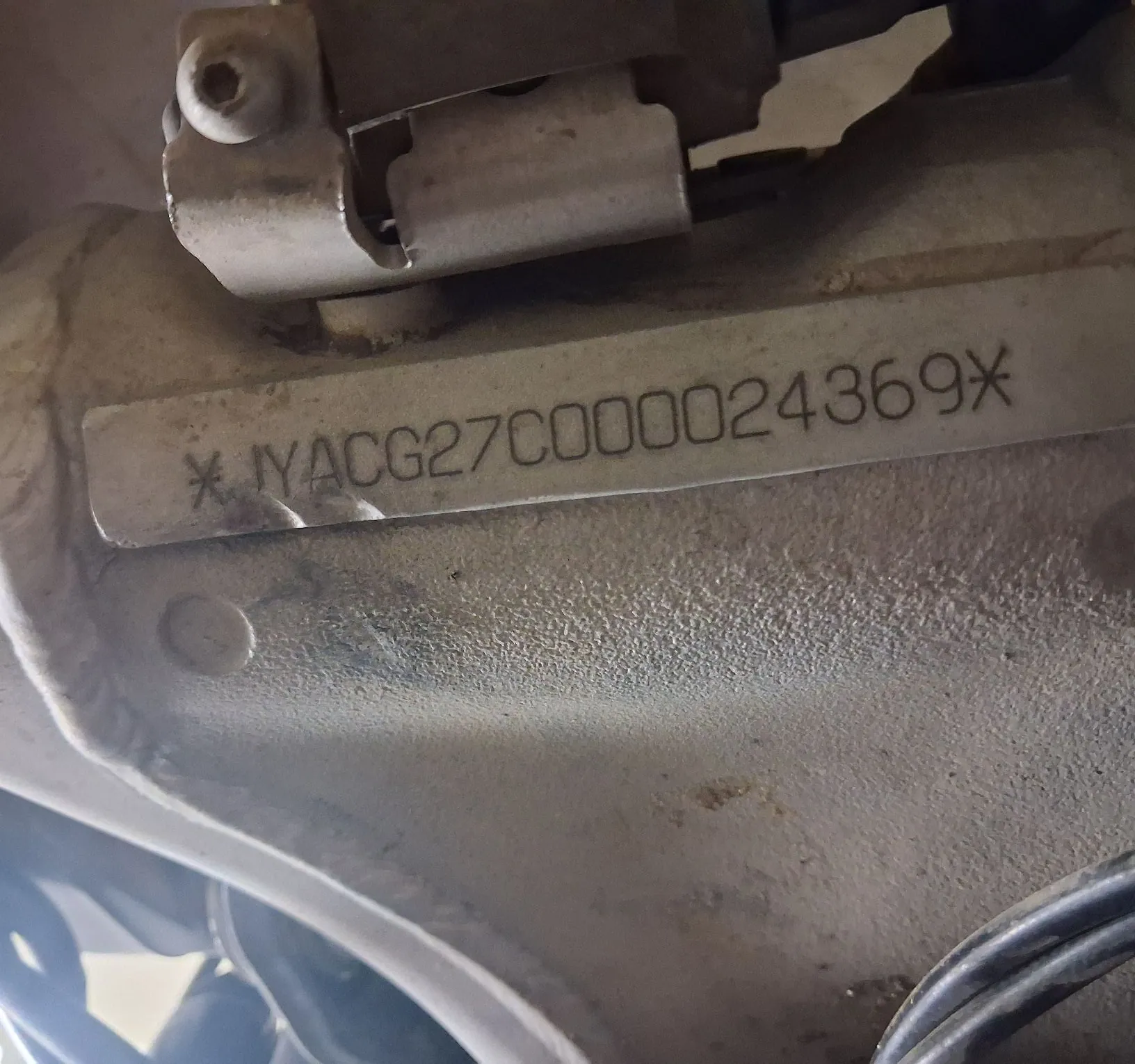Zbliżenie na metalowy element konstrukcyjny z przymocowaną tabliczką znamionową. Na tabliczce znajduje się pionowy napis: * YACG27C0000024369*.