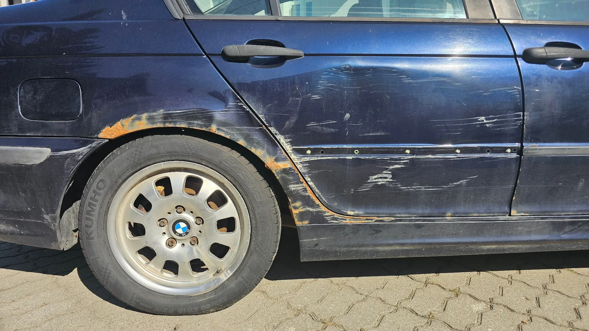 Zbliżenie na boczny panel ciemnoniebieskiego samochodu. Nad przednim lewym kołem widoczne są znaczne uszkodzenia karoserii w postaci rdzy oraz głębokich zarysowań i wgnieceń na drzwiach kierowcy. Samochód posiada srebrne felgi aluminiowe z logo BMW oraz opony marki Kumho.