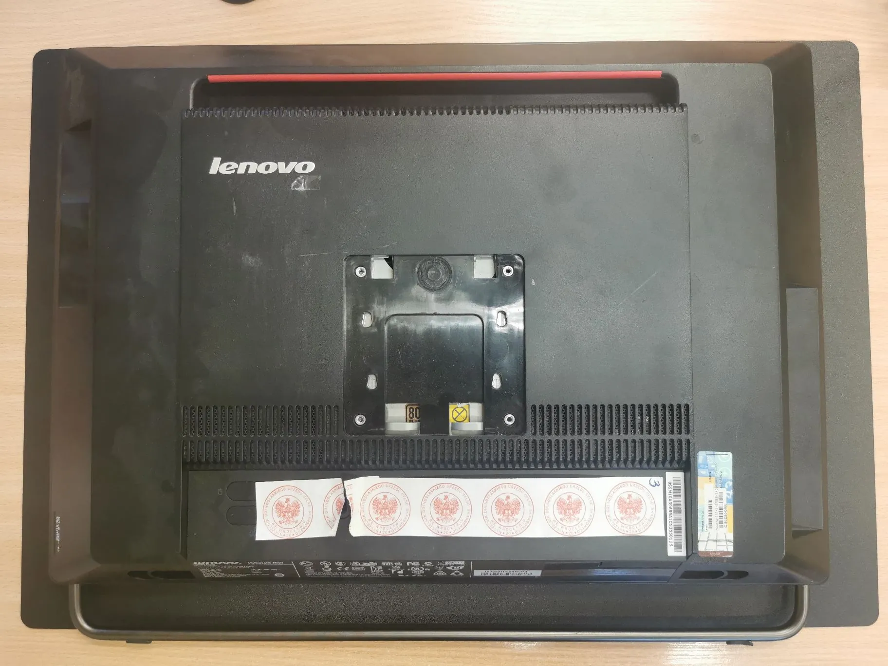 Zdjęcie przedstawiające tył czarnego monitora marki Lenovo. Na obudowie widoczne jest logo 'lenovo' w lewym górnym rogu. W centralnej części znajduje się metalowy uchwyt VESA do montażu na ścianie z czterema śrubami. Na dole obudowy przyklejone są białe naklejki z powtarzającym się czerwonym symbolem okrągłym oraz etykieta z kodem kreskowym i informacjami technicznymi. Na górze monitora widoczny jest czerwony element dekoracyjny.