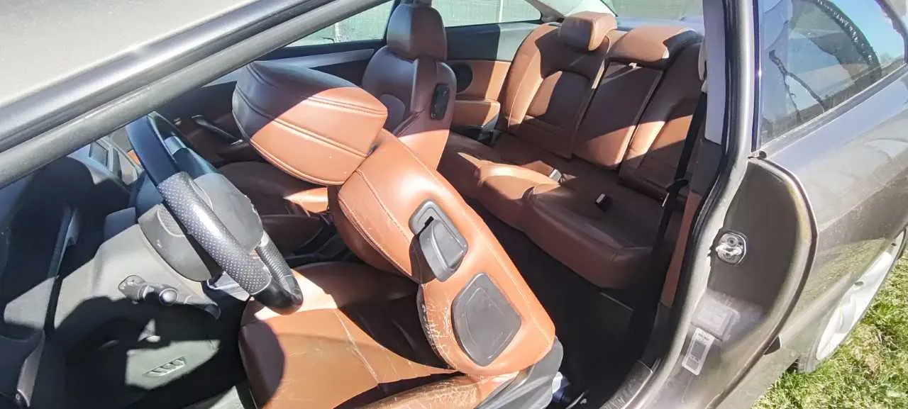 Wnętrze samochodu z otwartymi drzwiami od strony kierowcy. Widoczne brązowe skórzane fotele przednie i tylne, kierownica po lewej stronie, deska rozdzielcza oraz fragment zewnętrznej części pojazdu. Samochód zaparkowany na trawie, w tle elementy otoczenia. Brak czytelnego tekstu.