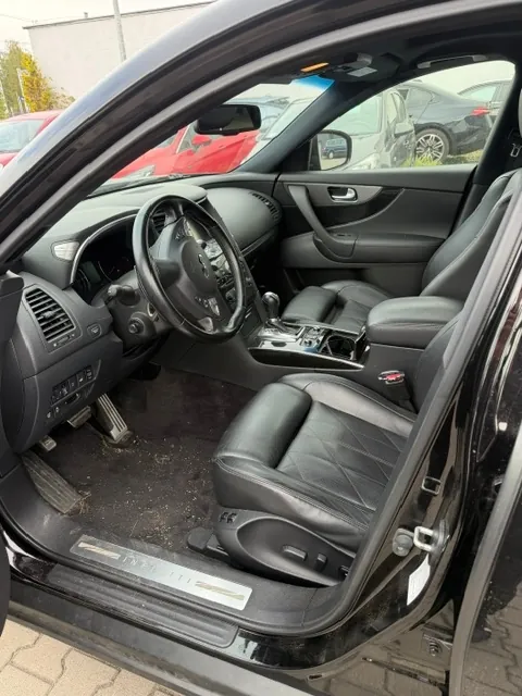 Wnętrze samochodu marki Infiniti widziane z otwartych drzwi kierowcy. Widoczny jest czarny skórzany fotel z przeszyciami w romby, kierownica, deska rozdzielcza z nawiewami oraz konsola środkowa. Na progu samochodu znajduje się metalowa listwa z napisem 'INFINITI'. Przez okna widać inne zaparkowane samochody w kolorach czerwonym i srebrnym.