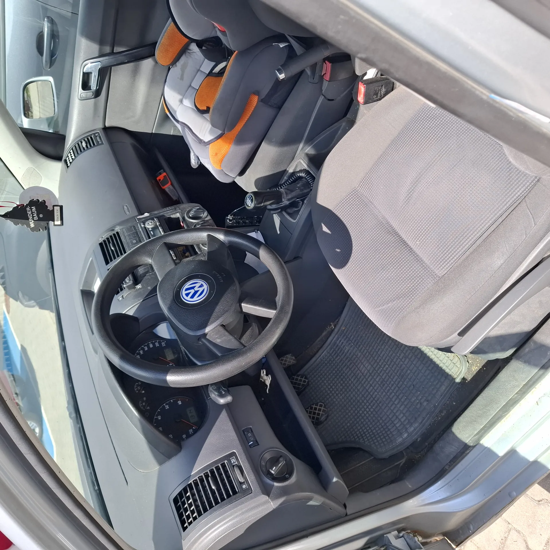 Wnętrze samochodu Volkswagen z widocznym kierownicą z logo marki, szarymi siedzeniami i fotelikiem dziecięcym z pomarańczowymi akcentami na tylnej kanapie. Na desce rozdzielczej widoczne są wskaźniki prędkości i obrotomierz, a na lusterku wstecznym zawieszony jest odświeżacz powietrza z napisem "WUNDER-BAUM". Drzwi pasażerskie są otwarte, a na podłodze leżą czarne maty. W tle widoczne są elementy kabiny, w tym dźwignia zmiany biegów i klimatyzacja.