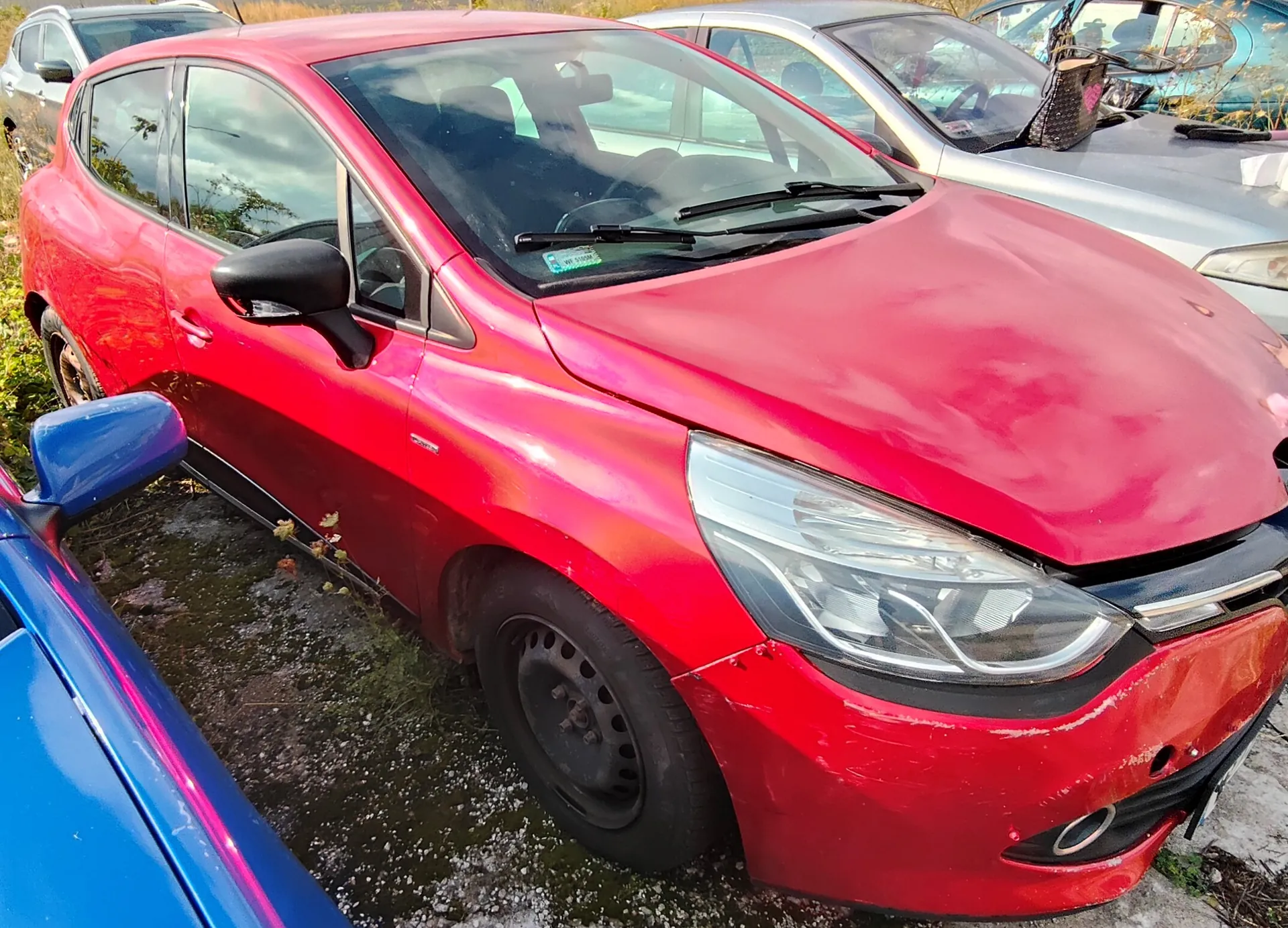 Czerwony samochód osobowy marki Peugeot w nadwoziu hatchback, zaparkowany na nieutwardzonym terenie obok innych pojazdów. Auto posiada czarne felgi stalowe i widoczne uszkodzenia lakieru na masce. W tle widoczne są fragmenty srebrnego i niebieskiego samochodu.