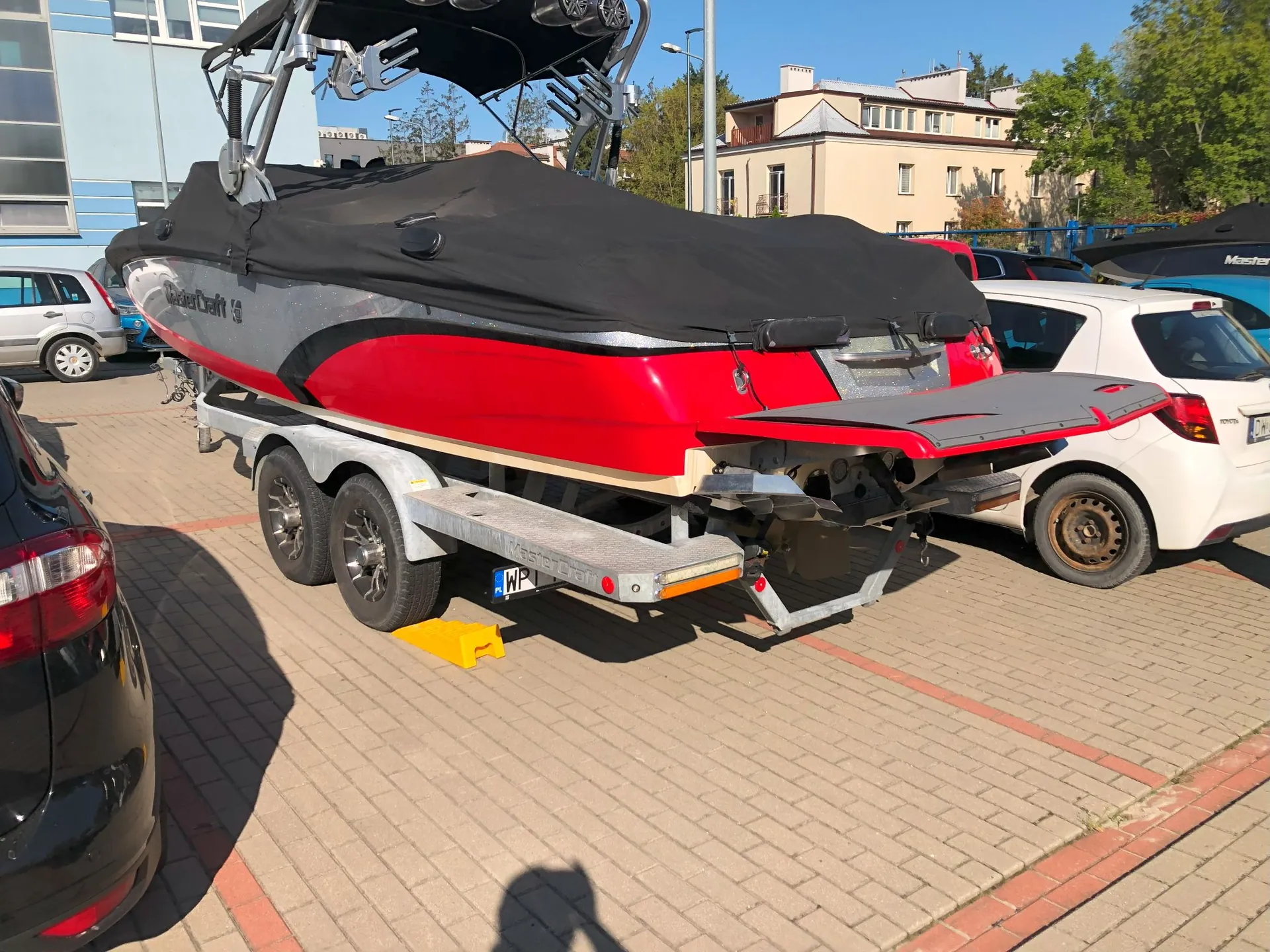Czerwono-biała łódź motorowa marki WaterCraft, przykryta czarną plandeką, zaparkowana na przyczepce na kostce brukowej. Obok łodzi stoją samochody: czarny po lewej i biały po prawej. W tle widoczny jest jasny budynek mieszkalny oraz niebo.