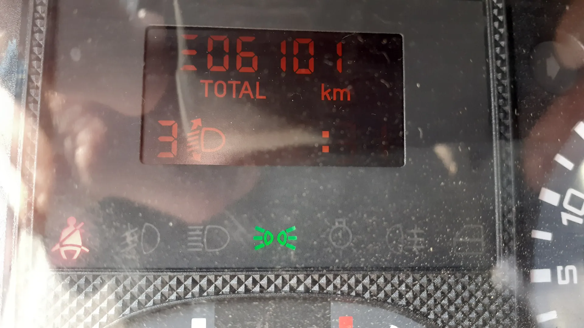 Zbliżenie na cyfrowy wyświetlacz samochodowy z czerwonymi cyframi 306101, poniżej napisem TOTAL i jednostką km. Poniżej widoczna cyfra 3 z symbolem przekładni oraz zielony symbol włączonych świateł. Na dole wyświetlacza umieszczone są ikony: czerwona ikona pasów bezpieczeństwa, szare symbole oraz zielony symbol świateł. Po prawej stronie częściowo widoczny jest analogowy wskaźnik prędkości z białymi znacznikami.