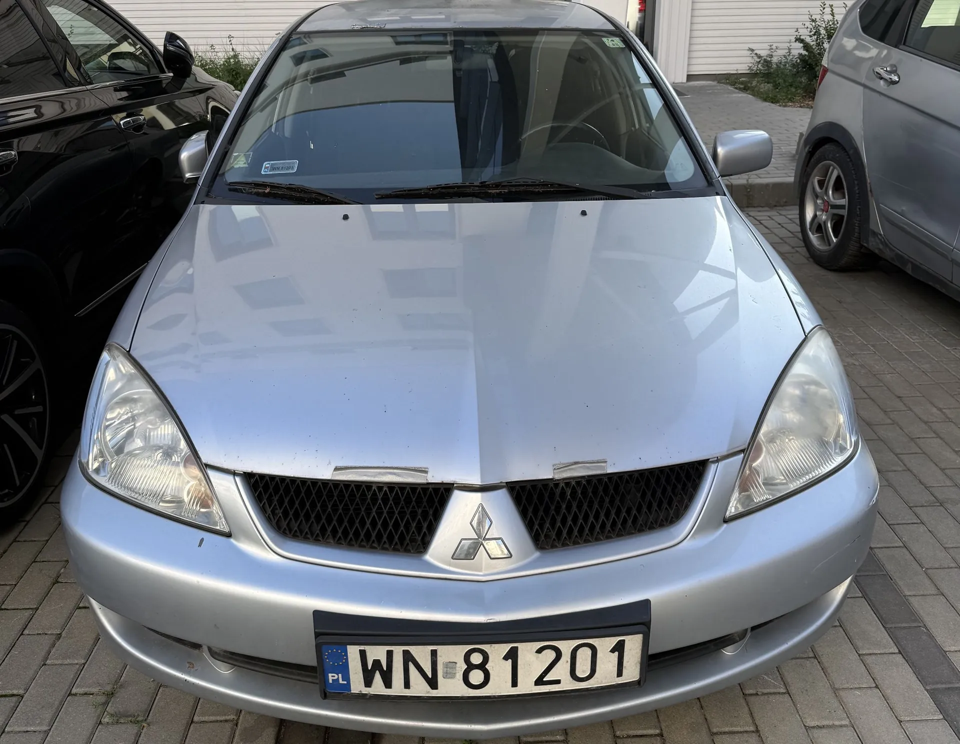 Srebrny samochód marki Mitsubishi widziany od przodu, zaparkowany na kostce brukowej. Na przednim zderzaku znajduje się polska tablica rejestracyjna o numerze WN 81201. W centralnej części grilla widoczne jest logo Mitsubishi.