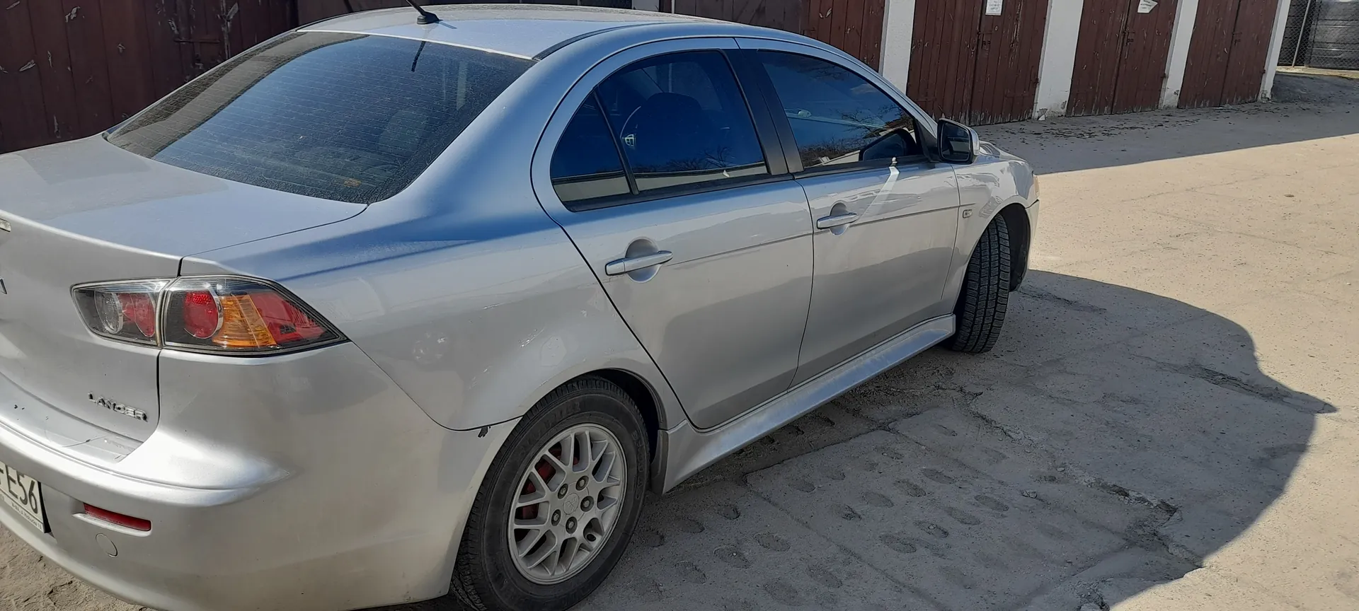 Srebrny samochód osobowy Mitsubishi Lancer zaparkowany na betonowej powierzchni z widocznymi odciskami stóp. Widok z tyłu i boku, z widocznym tylnym światłem, tablicą rejestracyjną z częścią numeru E56 oraz napisem LANCER na bagażniku. W tle brązowe drewniane ogrodzenie i białe filary budynku. Samochód stoi na płaskiej powierzchni, cień pada w prawo.