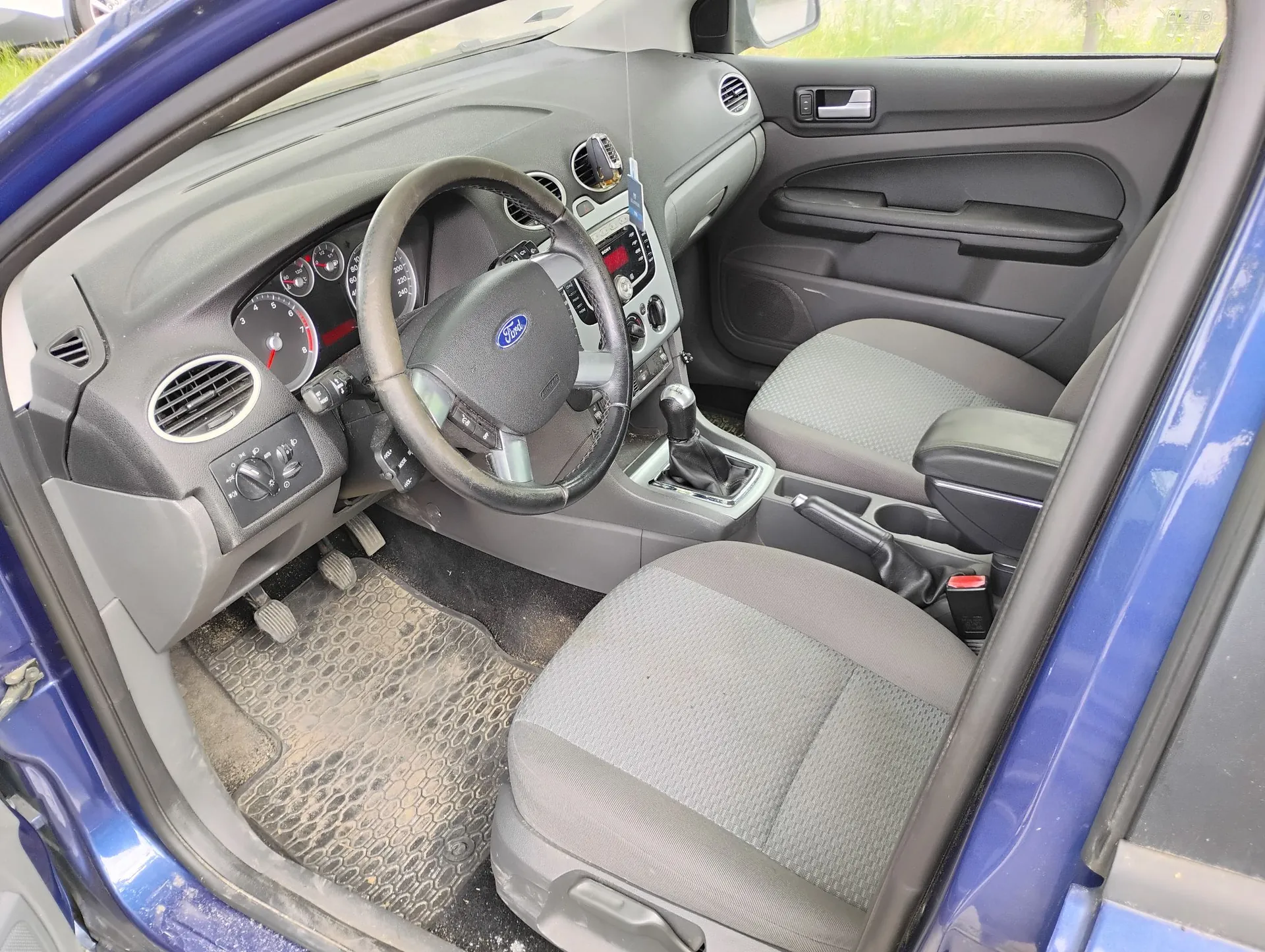 Wnętrze niebieskiego samochodu marki Ford, widok z otwartych drzwi pasażera. Widoczna jest szara deska rozdzielcza, kierownica z logo Ford, zegary, konsola środkowa z radiem, dwa szare materiałowe fotele oraz gumowa mata na podłodze.