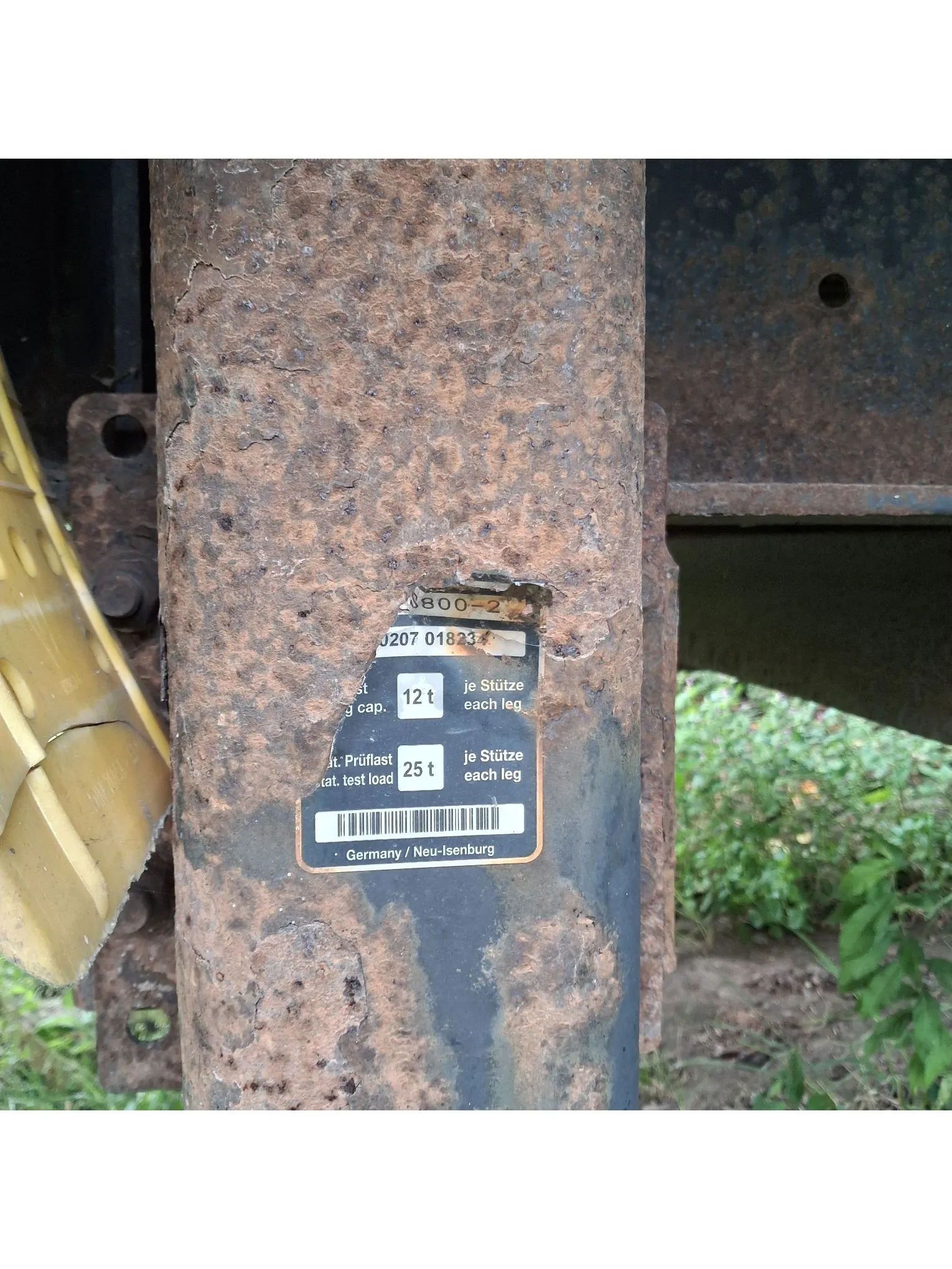 Zbliżenie na zardzewiały, metalowy element konstrukcyjny, prawdopodobnie część maszyny lub pojazdu. Na metalu znajduje się srebrna tabliczka znamionowa z tekstem w języku niemieckim i angielskim. Widoczne napisy to: '20800-2', '0207 018231', '12 t je Stütze each leg', 'Prüflast 25 t je Stütze test load each leg' oraz 'Germany / Neu-Isenburg'. Na dole tabliczki znajduje się kod kreskowy.