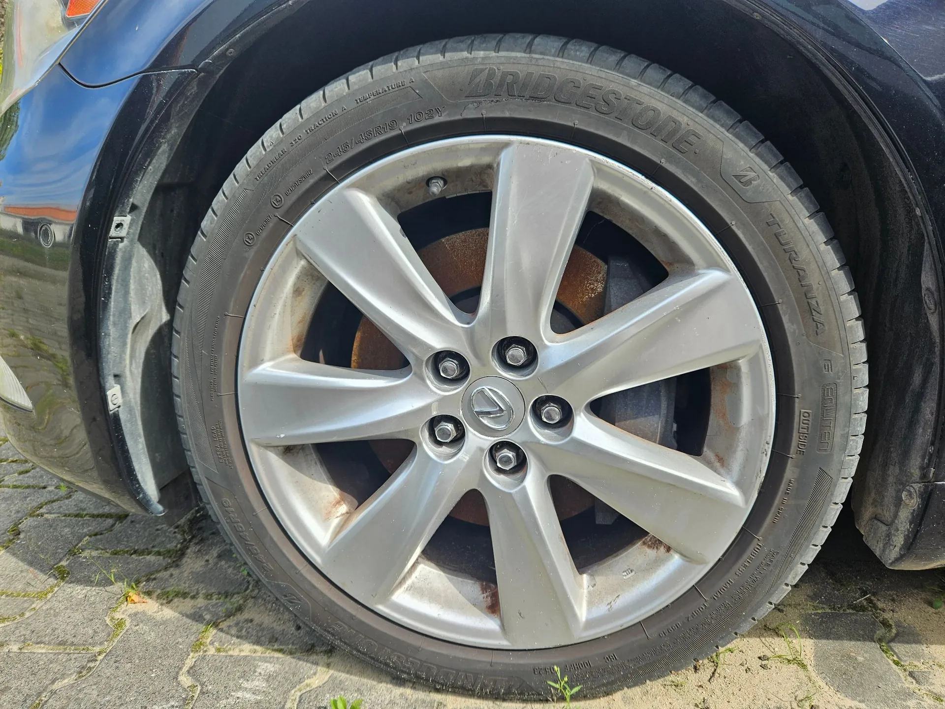 Zbliżenie na koło samochodu z aluminiową felgą o pięciu szerokich ramionach i oponą marki Bridgestone Turanza. Widoczny jest napis na oponie: 'BRIDGESTONE TURANZA'. Felga wykazuje oznaki zużycia i zabrudzenia, a w tle widoczny jest zardzewiały tarcza hamulcowa.