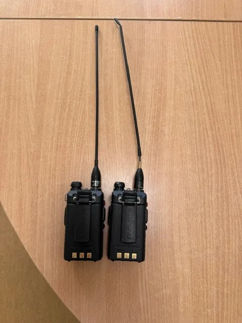 Dwie czarne radiostacje marki Baofeng leżące obok siebie na drewnianej powierzchni. Każde urządzenie posiada długą, pionową antenę. Na obudowach widoczny jest napis 'BAOFENG'.