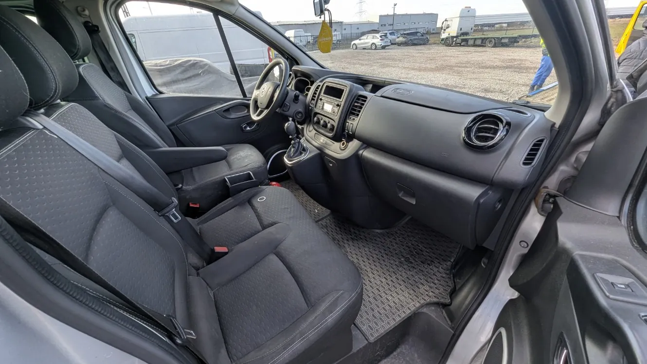 Wnętrze samochodu z widocznymi czarnymi siedzeniami o teksturze w kształcie kropek, kierownicą, deska rozdzielcza z przyciskami i ekranem, dźwignią zmiany biegów oraz dywanikami z gumową powierzchnią. Drzwi pasażerskie są otwarte, a na zewnątrz widoczne są inne pojazdy: biały furgon, samochody osobowe, ciężarówka oraz osoba w niebieskich spodniach na żwirowym placu. W tle budynki i maszty energetyczne.