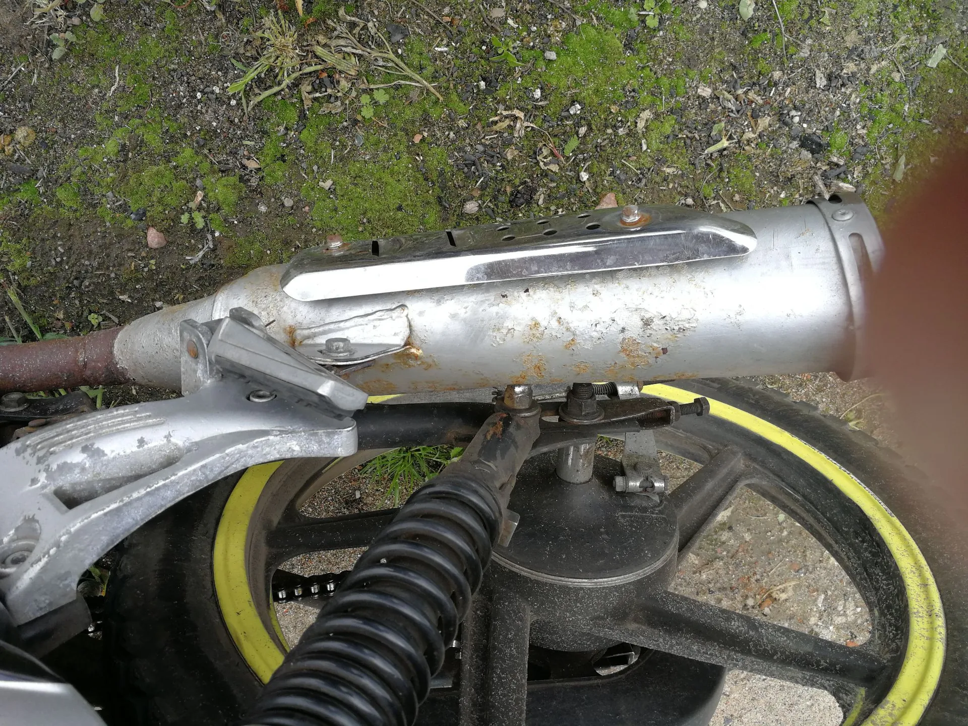 Zbliżenie na tylną część motocykla. Widoczny jest srebrny, metalowy tłumik z chromowaną osłoną przeciwszczypową, który wykazuje oznaki korozji. Poniżej znajduje się czarne koło z żółtą obwódką na oponie, amortyzator z czarną harmonijkową osłoną oraz srebrny element ramy lub podnóżka.