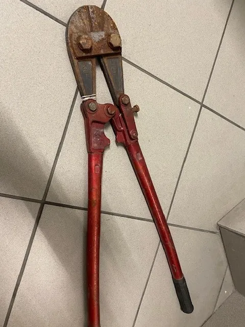 Czerwone nożyce do cięcia prętów z widoczną rdzą na części tnącej, leżące na białej podłodze z kafelkami o ciemnych szwach. Długa rękojeść z czarną osłonką na końcu jednej z rączek. Brak czytelnego tekstu.