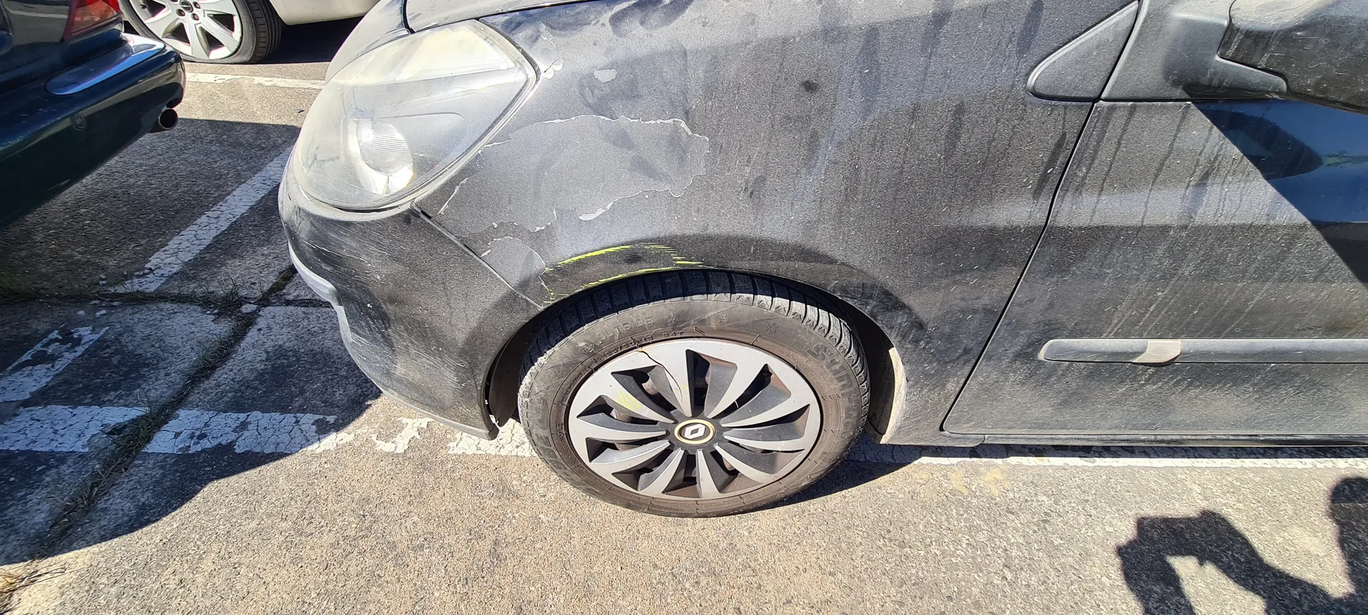 Czarny samochód zaparkowany na asfaltowym parkingu z białymi liniami. Widoczna przód pojazdu z uszkodzoną, obłupaną farbą na masce i błotniku. W centrum koła aluminiowego felga z logo Renault. W tle fragment innych samochodów oraz cień osoby na asfalcie. Samochód pokryty jest kurzem i plamami.