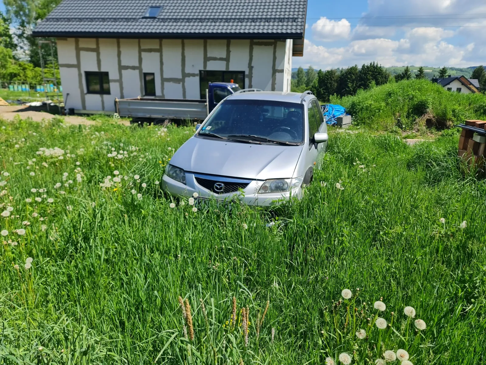 Srebrny samochód marki Mazda zaparkowany na wysokiej, zielonej trawie z białymi kwiatami mniszka. W tle budynek z białymi ścianami i ciemnym dachem, niebieski pojazd ciężarowy oraz zielone krzewy i drzewa. Niebo z białymi chmurami. Po prawej stronie widoczne są cegły i niebieska folia.