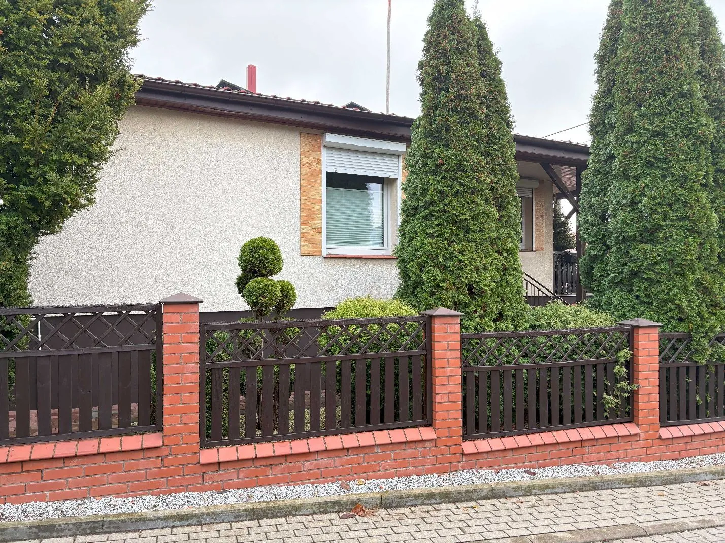 Zdjęcie jednokondygnacyjnego domu z jasną elewacją i ciemnym dachem. Przed domem znajduje się ogrodzenie wykonane z czerwonej cegły oraz brązowych drewnianych paneli z ozdobną kratką u góry. W ogrodzie rosną wysokie, smukłe tujy oraz mniejsze, formowane krzewy. W oknie domu widoczna jest opuszczona roleta zewnętrzna. Przed ogrodzeniem znajduje się chodnik z kostki brukowej.