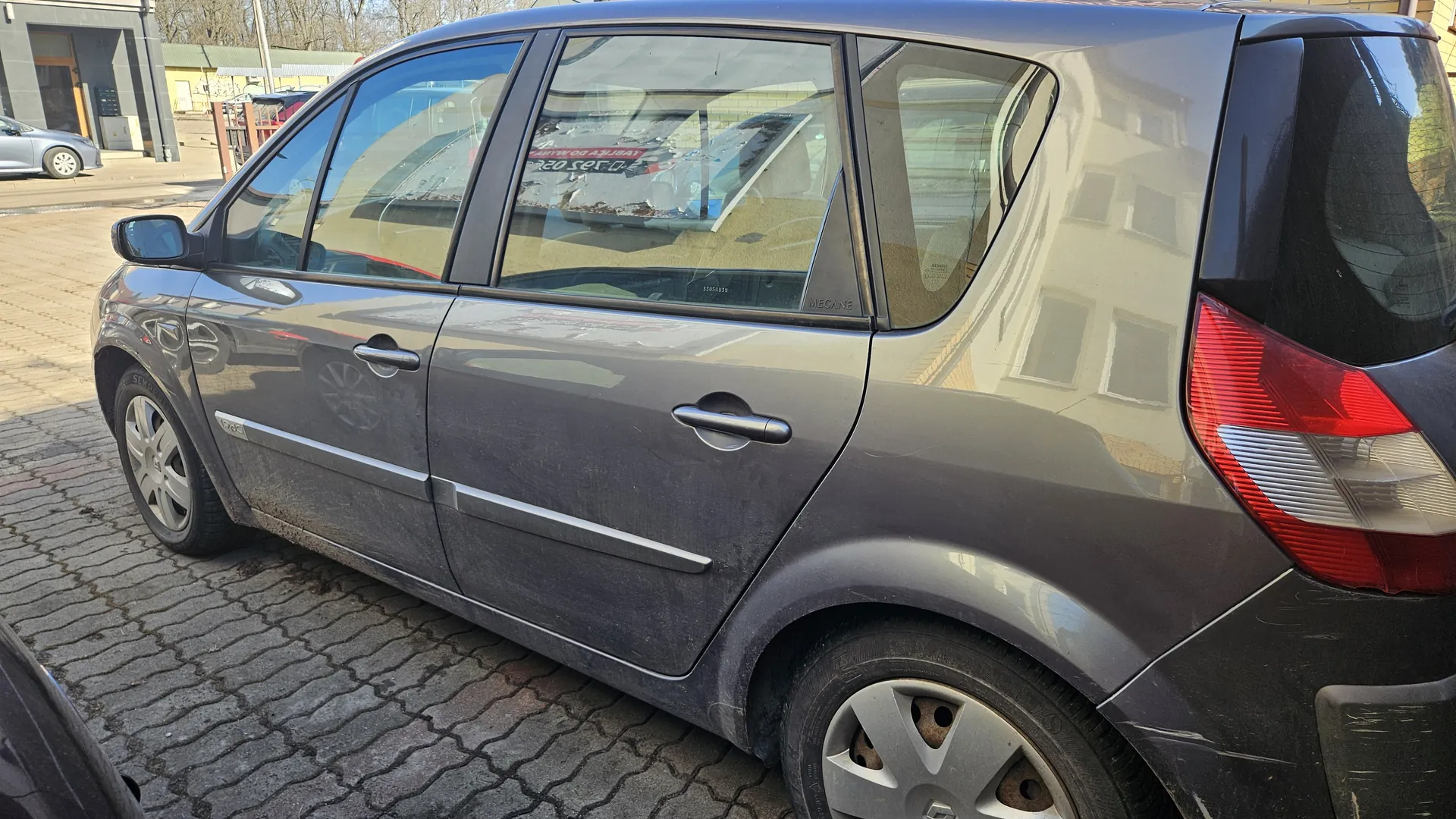 Szary samochód osobowy typu hatchback zaparkowany na brukowanej powierzchni. Widoczne są cztery drzwi, srebrne klamki, reflektory tylnego światła oraz odbicia budynków na karoserii. W tle stoją inne pojazdy i budynki z zielonym dachem. Na szybie bocznej widoczny napis 'MEGANE' oraz częściowo przyciemniony napis w języku polskim. Samochód pokryty jest kurzem, a na felgach widoczne są ślady brudu.