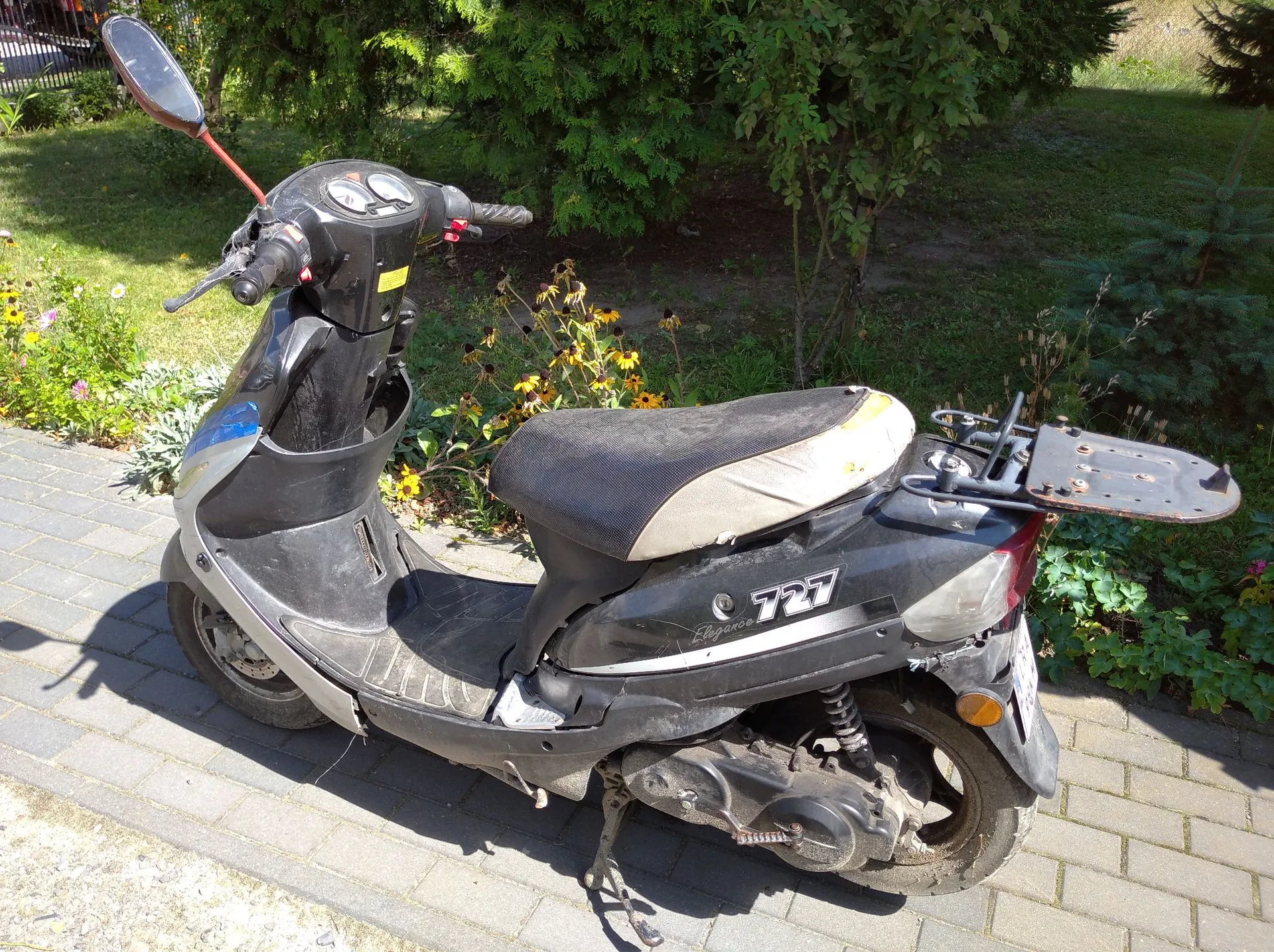 Czarny skuter zaparkowany na kostce brukowej przed zielonymi krzewami. Pojazd ma czarne nadwozie z napisem 'Elegance 125' na boku, czarno-szare siedzenie oraz lusterko po lewej stronie. Skuter stoi na bocznej stopce.