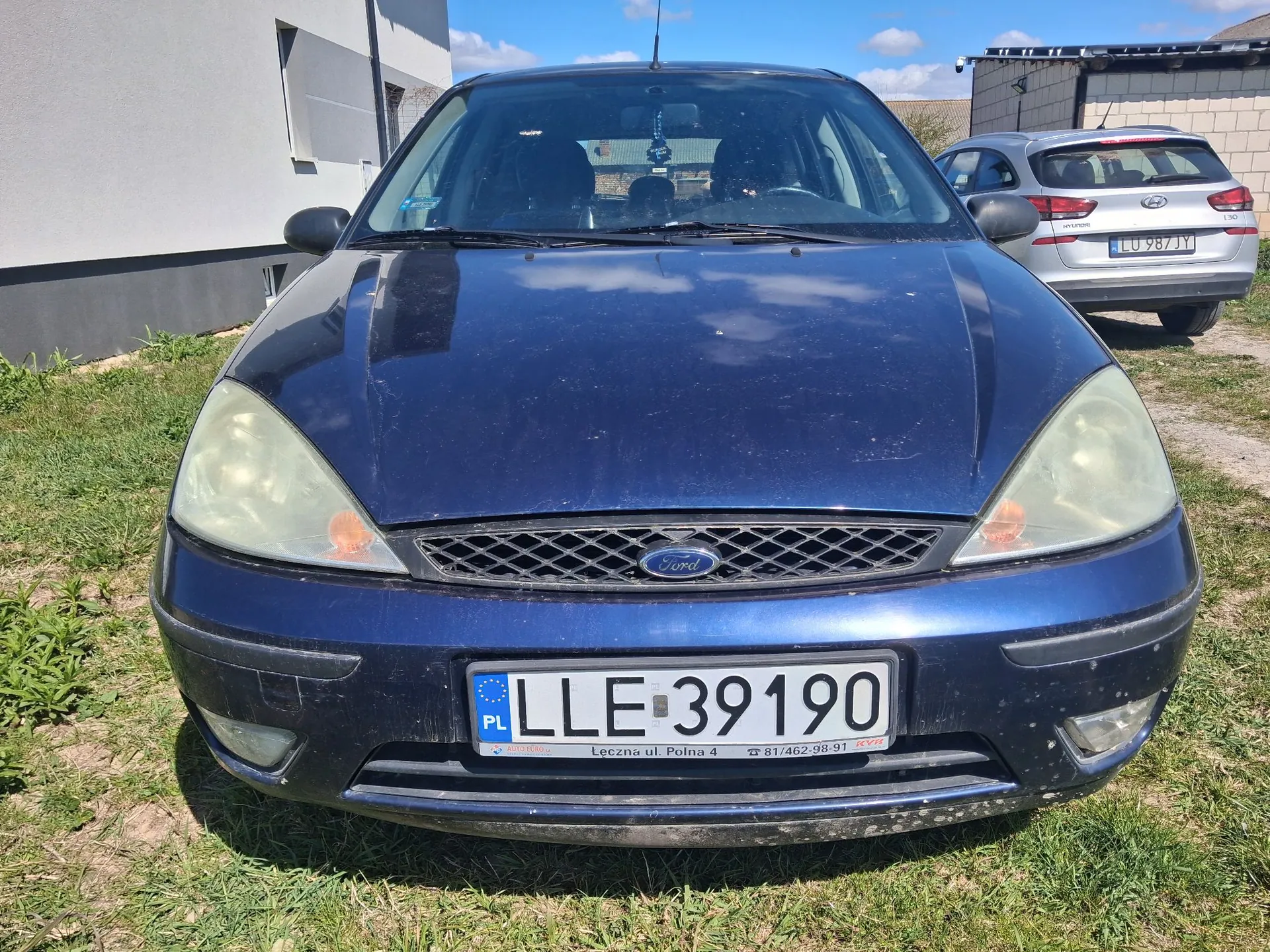 Widok z przodu ciemnoniebieskiego samochodu marki Ford zaparkowanego na trawie. Auto ma zmatowiałe reflektory i polską tablicę rejestracyjną o numerze LLE 39190. Poniżej numeru rejestracyjnego widnieje napis: 'Leczna ul. Polna 4 81/462 98-91'. W tle po prawej stronie widoczny jest tył srebrnego samochodu marki Hyundai z tablicą rejestracyjną LU 987JY oraz fragmenty budynków pod niebieskim niebem.