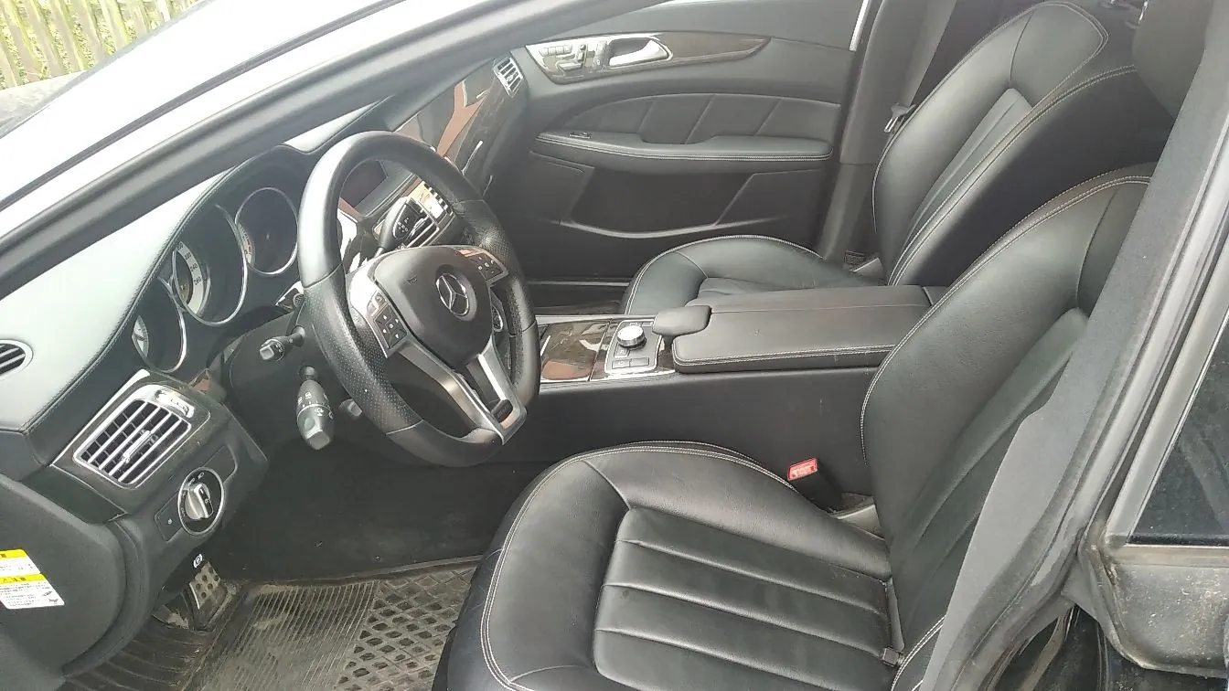 Wnętrze samochodu marki Mercedes-Benz z czarną skórzaną tapicerką. Widoczna jest kierownica z logo marki, deska rozdzielcza z zegarami, konsola środkowa z podłokietnikiem oraz dwa przednie fotele.