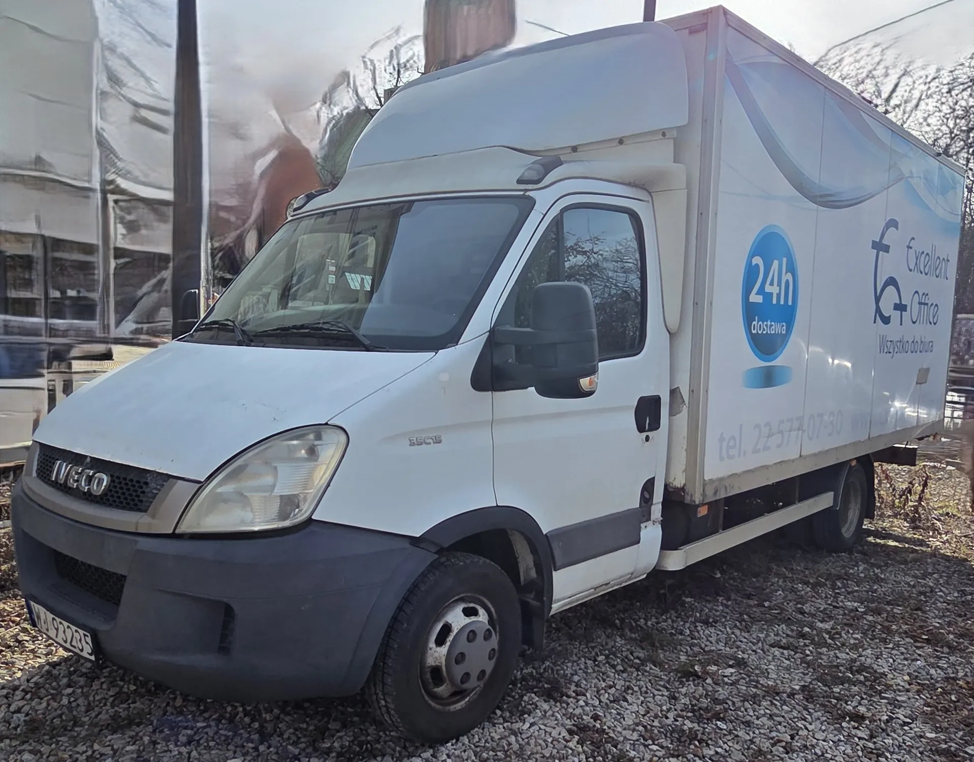 Biały samochód dostawczy marki Iveco zaparkowany na żwirowym podłożu. Na boku pojazdu widoczne są napisy: niebieski okrąg z białym tekstem '24h dostawa', logo 'Excellent Office' oraz 'Wszystko do biura'. Poniżej znajduje się numer telefonu 'tel. 22-577-07-30' i fragment adresu strony internetowej. Na przodzie samochodu widoczny jest napis 'IVECO' oraz tablica rejestracyjna 'WJ 93235'. W tle budynki, drzewa i częściowo widoczny inny pojazd.