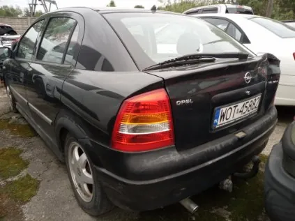 Czarny samochód marki Opel widziany od tyłu, z tablicą rejestracyjną WOT-4588. Pojazd stoi na podłożu z żwirem i trawą, w tle widoczne są inne samochody oraz zalesiony teren. Na tylnym szybie i bagażniku widoczne są elementy konstrukcyjne i logo marki.