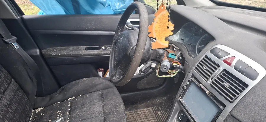 Wnętrze samochodu z widocznym kierownicą po lewej stronie, brudnymi czarnymi siedzeniami pokrytymi drobnymi śmieciem, uszkodzonym deskiem rozdzielczą z odsłoniętymi przewodami i brakującymi elementami. Na kierownicy wiszą pomarańczowy odświeżacz powietrza w kształcie liścia. Widoczne są zanieczyszczone drzwi pasażera, klimatyzacja oraz ekran centralny. Za oknem widoczna niebieska folia. Całość w stanie zaniedbania.