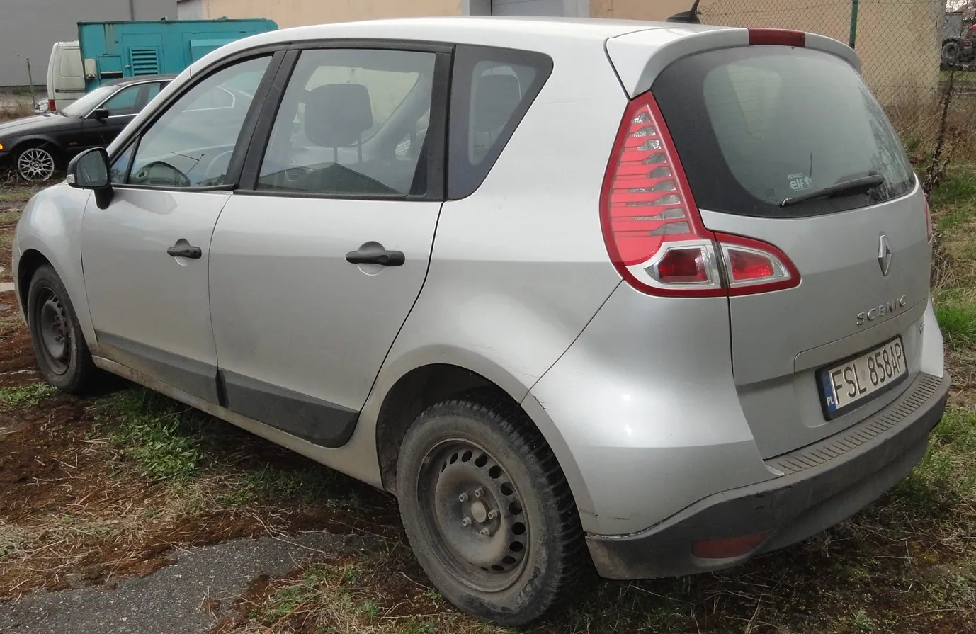 Srebrny samochód Renault Scenic zaparkowany na trawiastym i błotnistym terenie. Widoczna tylna część pojazdu z czerwonymi światłami, tablicą rejestracyjną FSL 858AP oraz napisem SCENIC na bagażniku. W tle czarny samochód, niebieski kontener i ogrodzenie. Opony mają brudne felgi, a nadkola są częściowo zabrudzone.