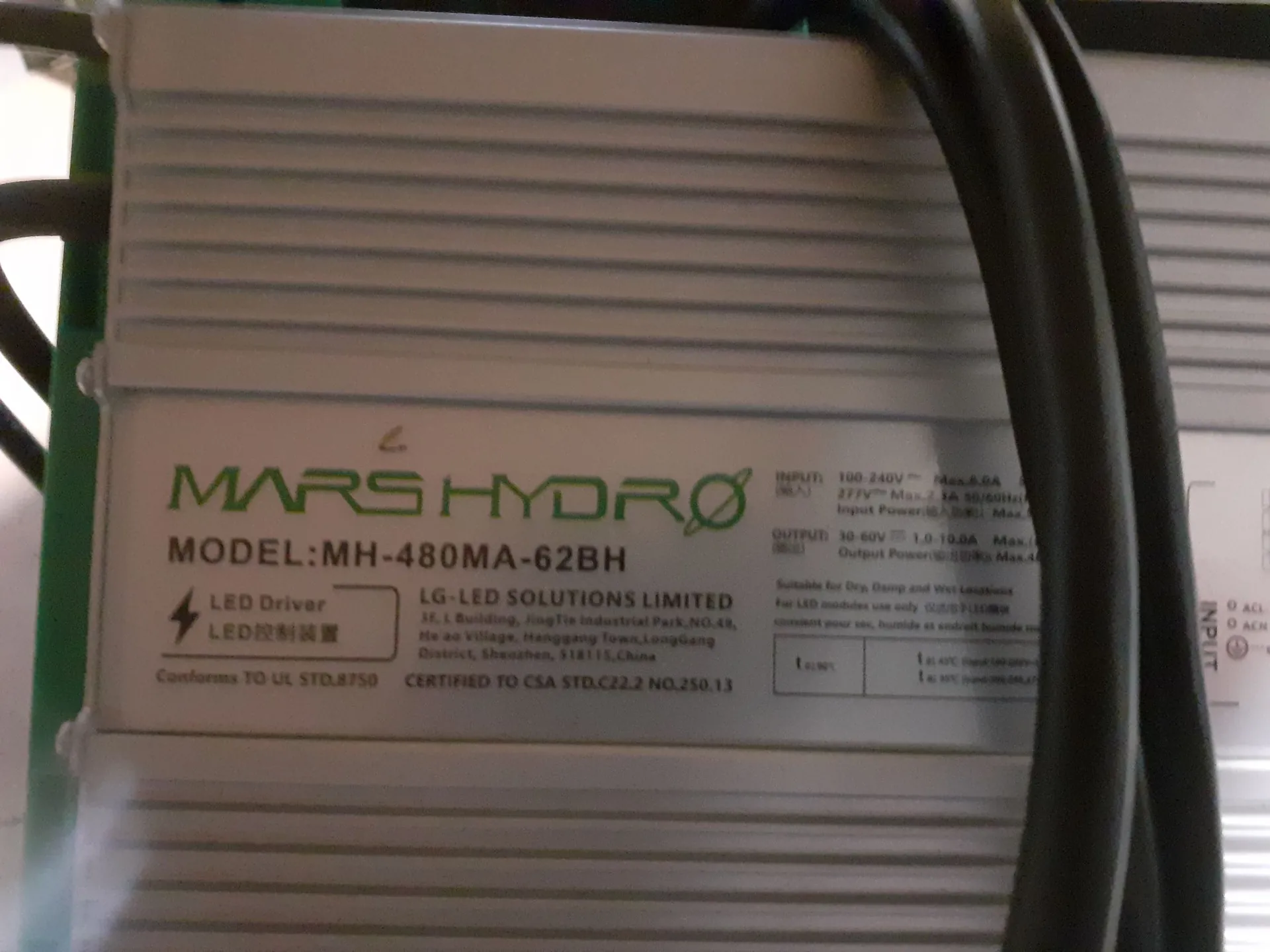 Zbliżenie na srebrną, aluminiową obudowę zasilacza LED z naklejką informacyjną. Na naklejce widnieje logo 'MARS HYDRO' oraz napis 'MODEL: MH-480MA-62BH'. Poniżej znajduje się tekst 'LED Driver' oraz dane producenta: 'LG-LED SOLUTIONS LIMITED, 3F, L Building, JingTie Industrial Park, NO.48, He ao Village, Hanggang Town, LongGang District, Shenzhen, 518115, China'. Widoczne są również specyfikacje techniczne dotyczące wejścia (INPUT: 100-240V) i wyjścia (OUTPUT: 30-60V), a także certyfikaty 'Conforms TO UL STD.8750' i 'CERTIFIED TO CSA STD.C22.2 NO.250.13'. Po prawej stronie widoczne są czarne kable zasilające.