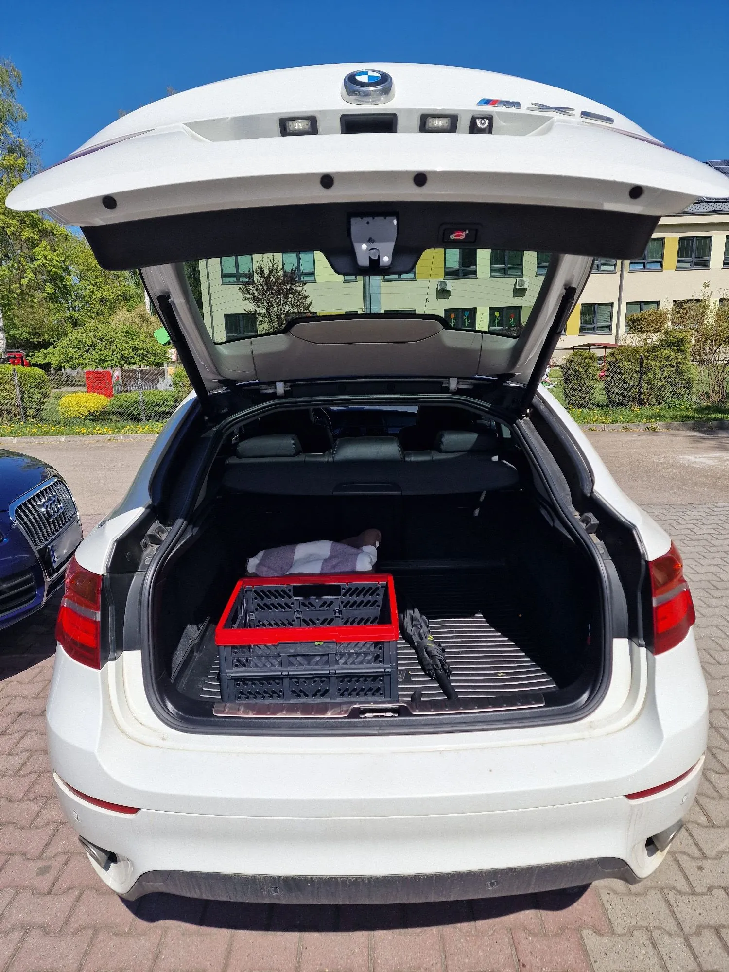 Widok z tyłu białego samochodu marki BMW z otwartą klapą bagażnika. Wewnątrz bagażnika znajduje się czarna plastikowa skrzynka z czerwonymi krawędziami oraz jasny przedmiot przypominający poduszkę lub zwinięty koc. W tle widoczny jest budynek oraz drzewa pod bezchmurnym, niebieskim niebem.