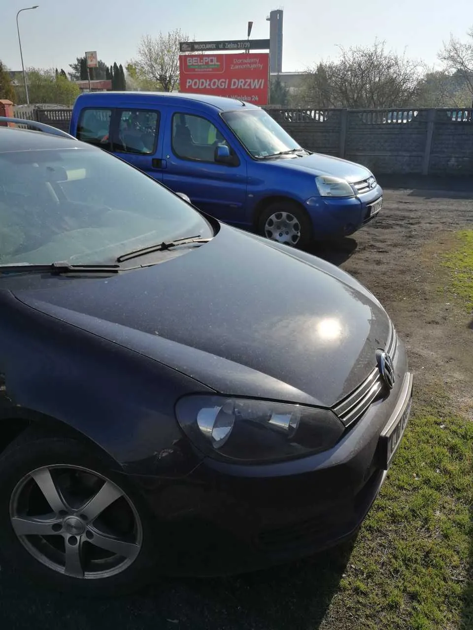Dwa samochody zaparkowane na otwartym obszarze: na pierwszym planie ciemnoszary samochód osobowy marki Volkswagen z widocznym logo, na drugim planie niebieski samochód dostawczy. W tle czerwony szyld z napisami 'BEL-POL', 'PODŁOGI DRZWI' oraz mniejszym tekstem 'Doradzamy Zamienniki'. Za samochodami betonowy płot, drzewa bez liści, słup telekomunikacyjny i budynek. Podłoga składa się z trawy i brudnej nawierzchni. Niebo jest jasne.