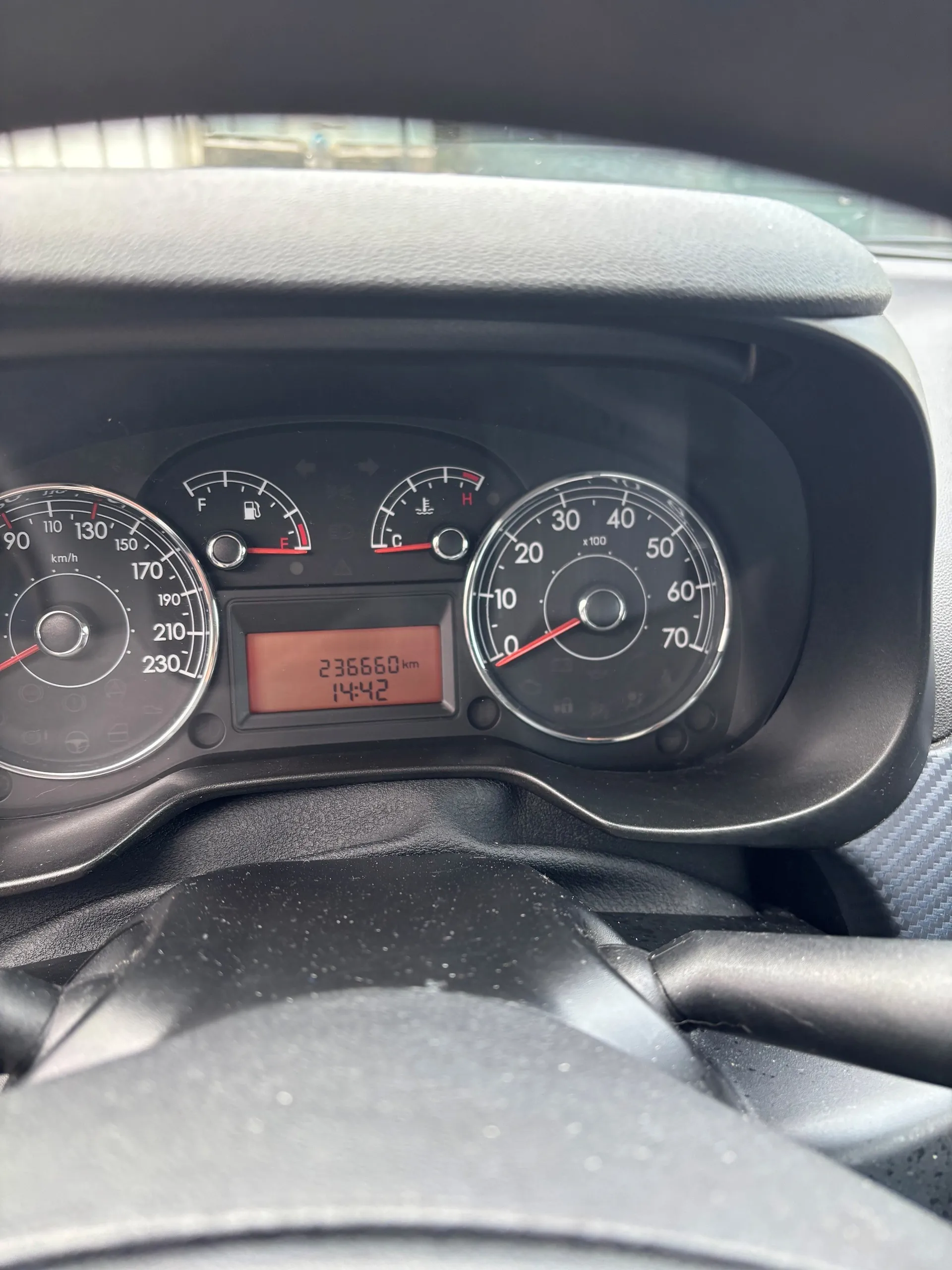 Zbliżenie na deski rozdzielcze samochodu. Widoczne są trzy analogowe wskaźniki: po lewej prędkościomierz z zakresem do 230 km/h, w środku wskaźnik paliwa (F/E) i temperatury chłodniczego (C/H), po prawej obrotomierz z zakresem do 70 (x100). W centrum wyświetlacz cyfrowy z przebiegiem 236660 km i godziną 14:42. Na dole widoczna część kierownicy. Tło stanowi fragment przedniej szyby i deski rozdzielczej.