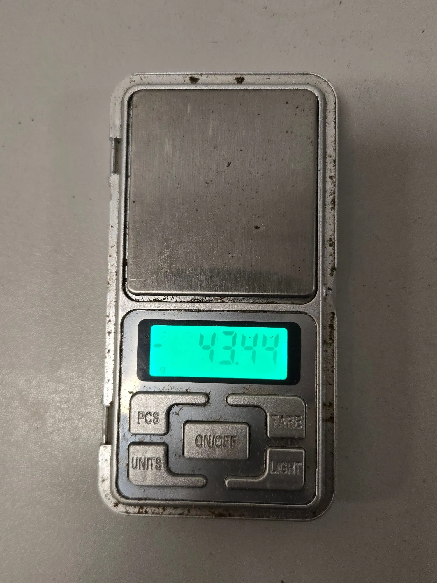 Srebrna, cyfrowa waga kieszonkowa z metalową szalką pomiarową. Wyświetlacz LCD świeci na zielono i pokazuje wartość 43.44. Poniżej wyświetlacza znajdują się cztery przyciski z napisami: PCS, TARE, UNITS oraz LIGHT, a w centrum większy przycisk ON/OFF. Obudowa wagi jest zabrudzona.