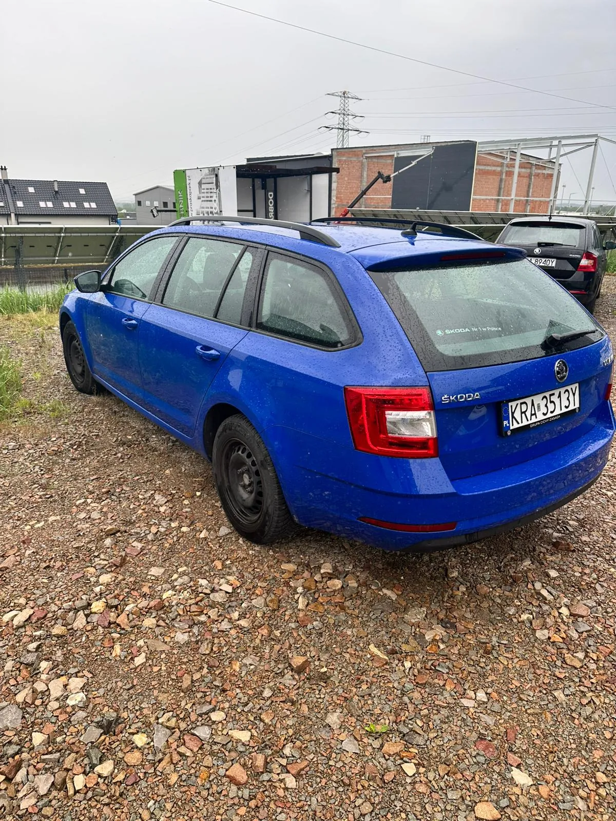 Niebieski samochód osobowy typu kombi marki ŠKODA zaparkowany na żwirowym podłożu. Widoczny tył pojazdu z tablicą rejestracyjną KRA 3513Y oraz napisem ŠKODA na bagażniku. W tle czarny samochód z tablicą 8940Y, budynki przemysłowe, słup energetyczny i zielona tablica reklamowa. Niebo jest zachmurzone, a na powierzchni samochodu widoczne są krople deszczu.