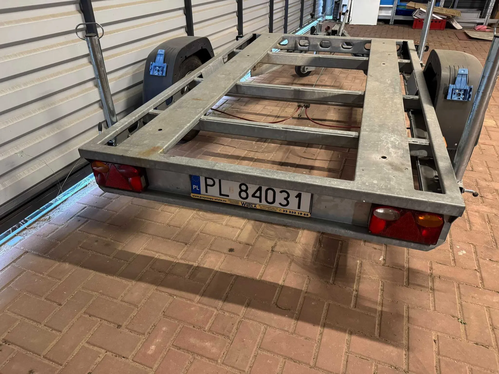 Metalowa rama przyczepy zaparkowana na kostce brukowej przed ścianą z blachy falistej. Rama jest pusta, widoczne są koła oraz tablica rejestracyjna o treści 'PL 84031'. Poniżej tablicy znajduje się żółty pasek z tekstem 'www.przyczepy-wola.eu Wola' oraz numerem telefonu '22 332 19 65'. Po bokach ramy znajdują się czerwone światła odblaskowe.