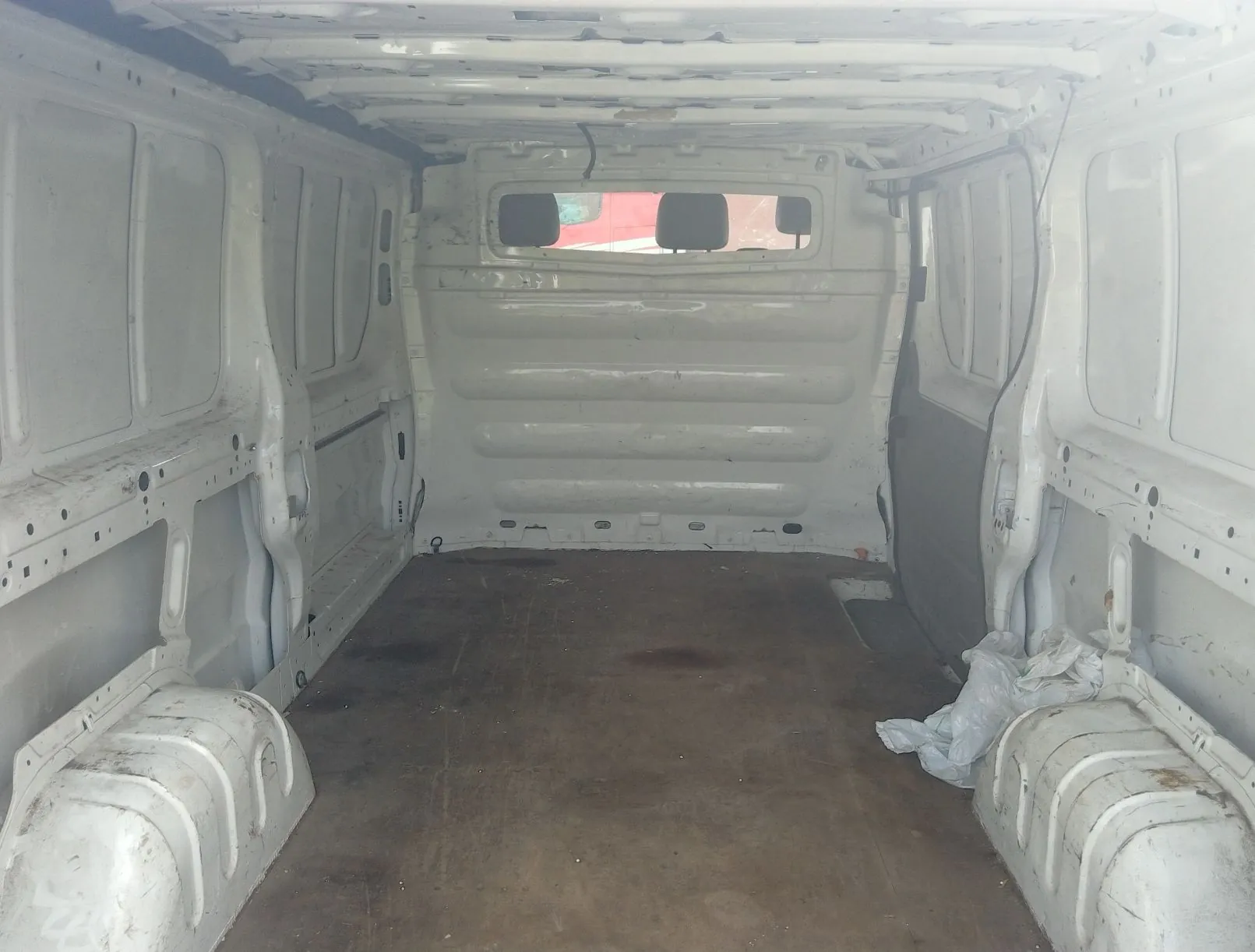 Wnętrze pustej przestrzeni ładunkowej białego samochodu dostawczego. Widoczna jest drewniana podłoga, białe metalowe ściany boczne oraz tylna ścianka z małym okienkiem. Po prawej stronie na podłodze leży zwinięta biała folia plastikowa.