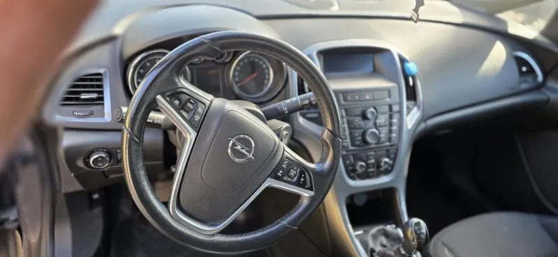 Wnętrze samochodu z kierownicą z logo Opel, tablicą przyrządów z dwoma okrągłymi wskaźnikami, centralnym panelem sterowania z przyciskami i pokrętłami oraz dźwignią zmiany biegów. Widoczne są również fragmenty deski rozdzielczej i siedzenia kierowcy.